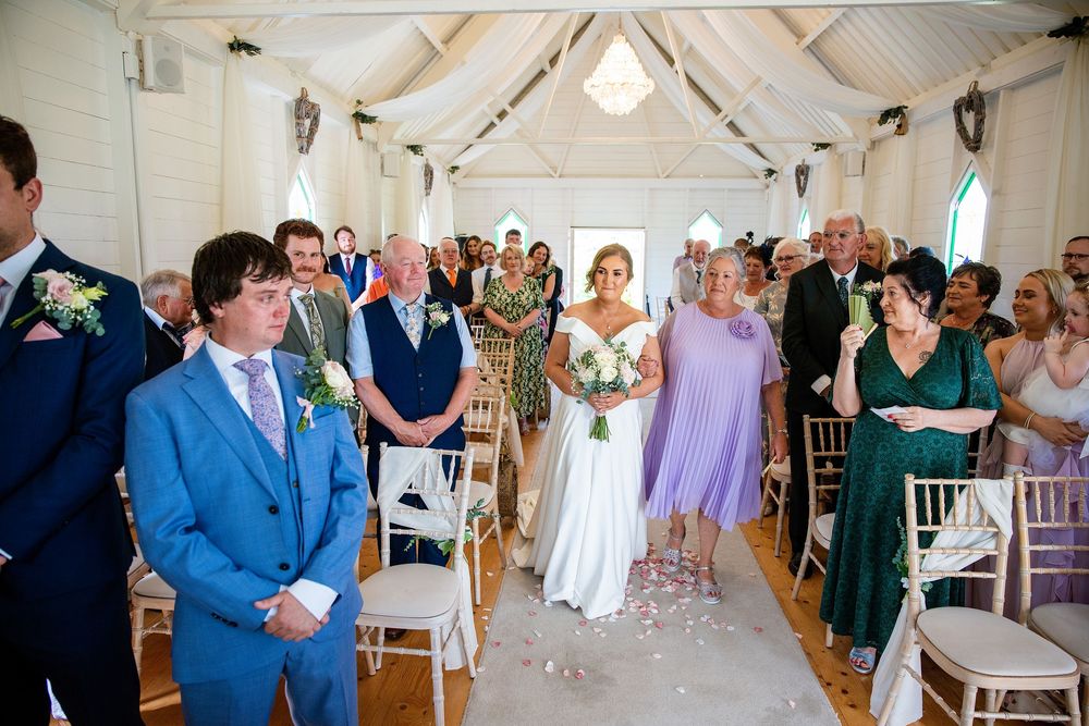 Bride walks down the aisle in white gown at elegant indoor wedding ceremony with guests standing.