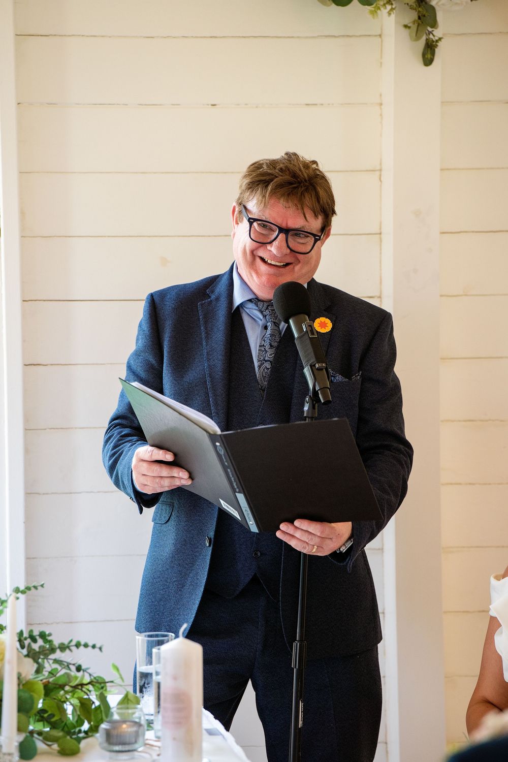 Man in navy suit with orange boutonniere smiles while reading from folder at wedding ceremony microphone.