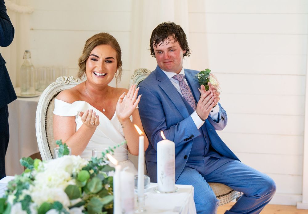 Bride and groom clapping hands joyfully at wedding ceremony, seated at decorated table with candles and greenery.