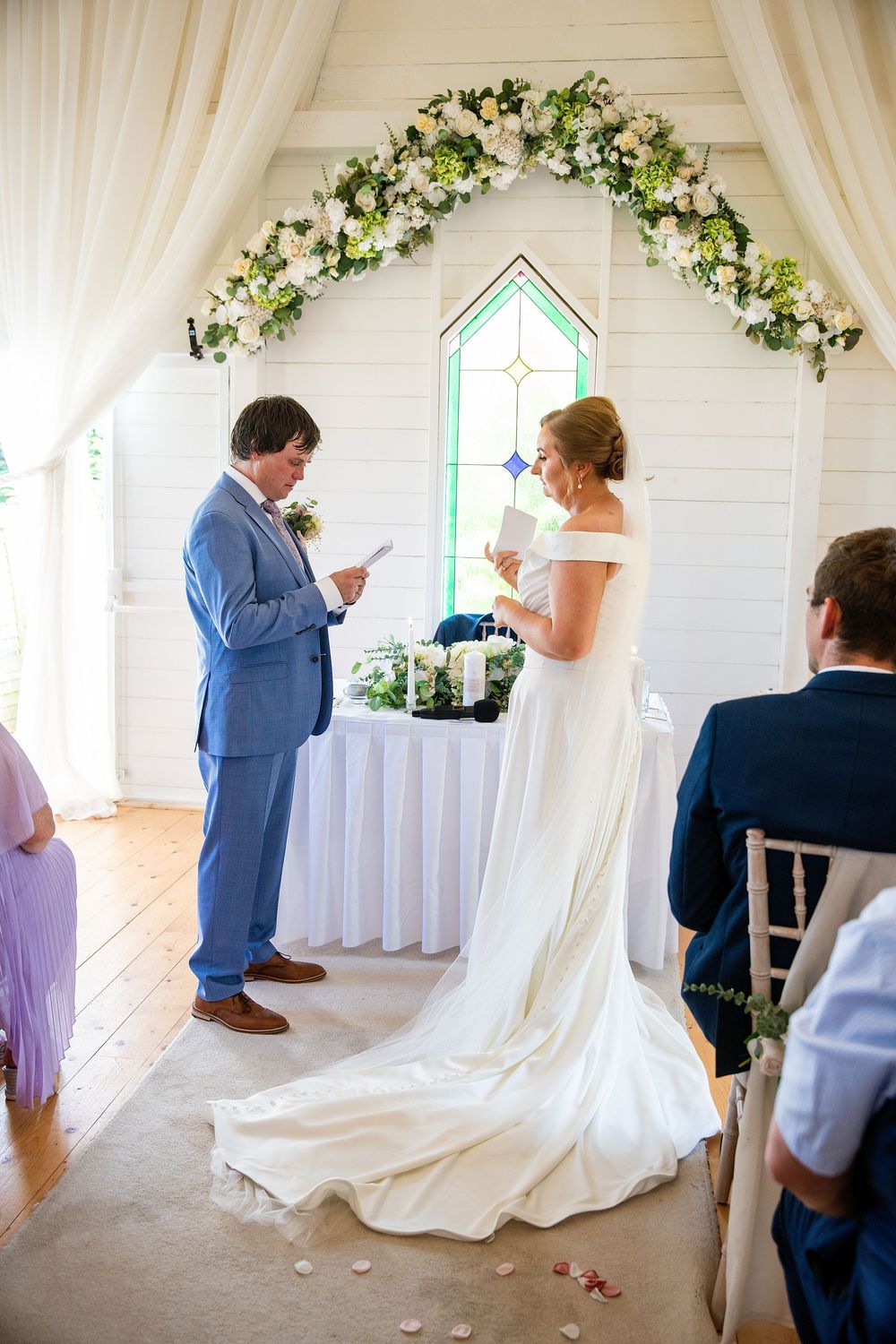 Bride and groom exchange vows in a white chapel adorned with a floral arch of white flowers and greenery.