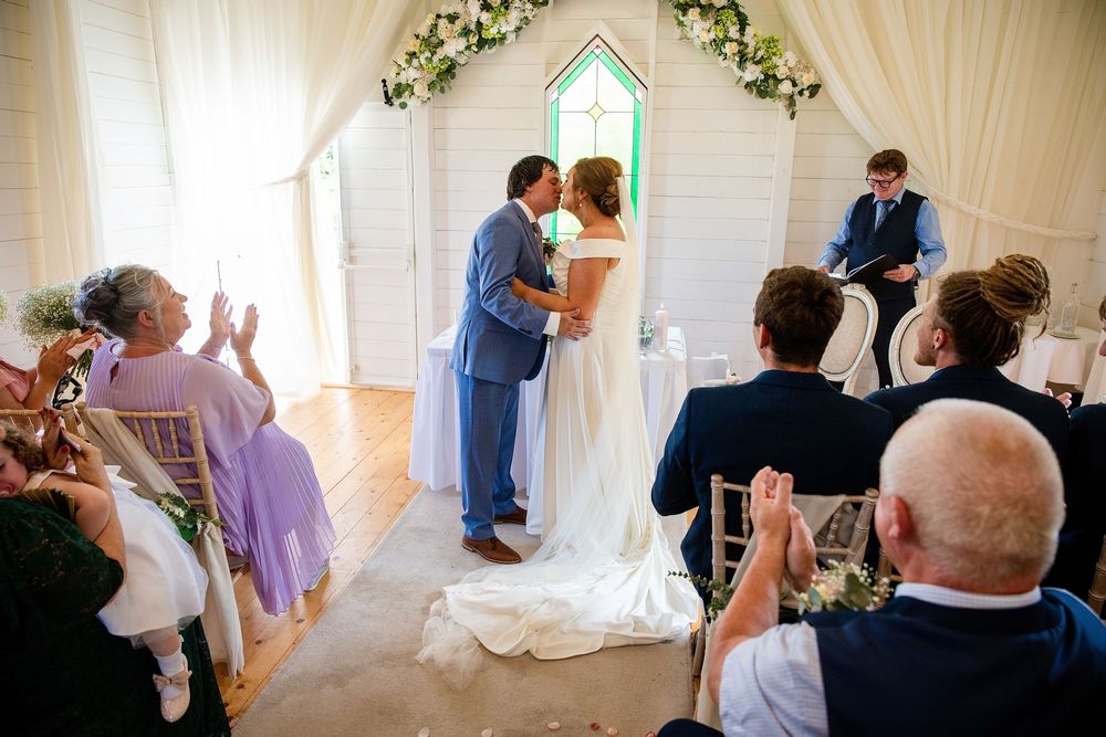 Couple shares first kiss at wedding altar in white chapel while guests applaud and photographer captures the moment.