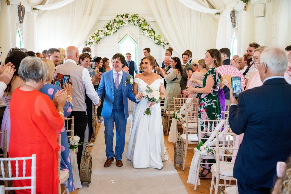 Bride and groom walk down the aisle at an elegant wedding ceremony venue with floral arch and guests applauding.