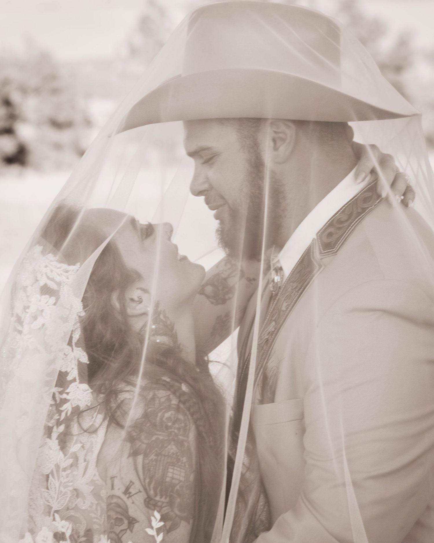 Black and white photo of a bride and groom sharing a romantic moment under a veil, groom wearing a cowboy hat.