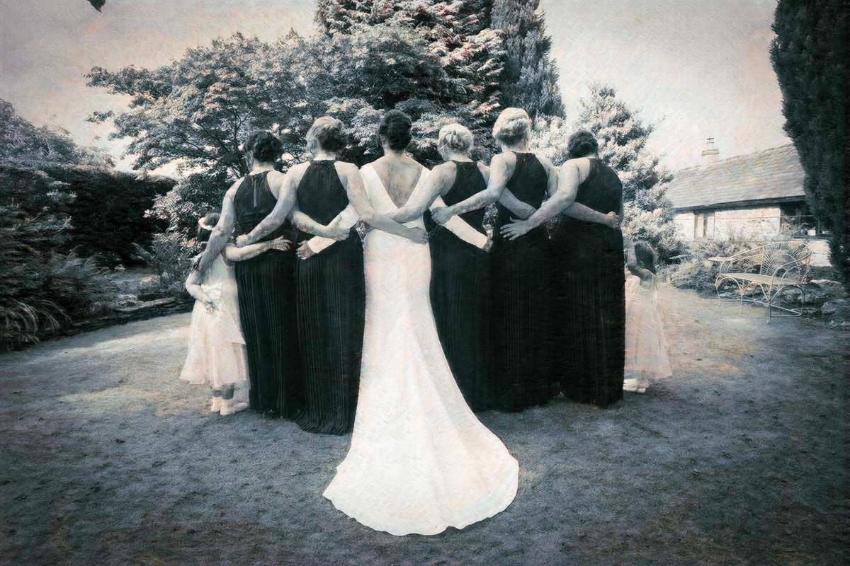Bride in white gown stands with bridesmaids in dark dresses, backs to camera, in a garden setting.