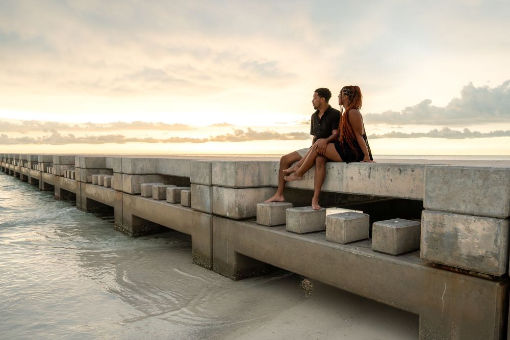 Couple sitting on a concrete pier at sunset overlooking a calm ocean with a dramatic cloudy sky.