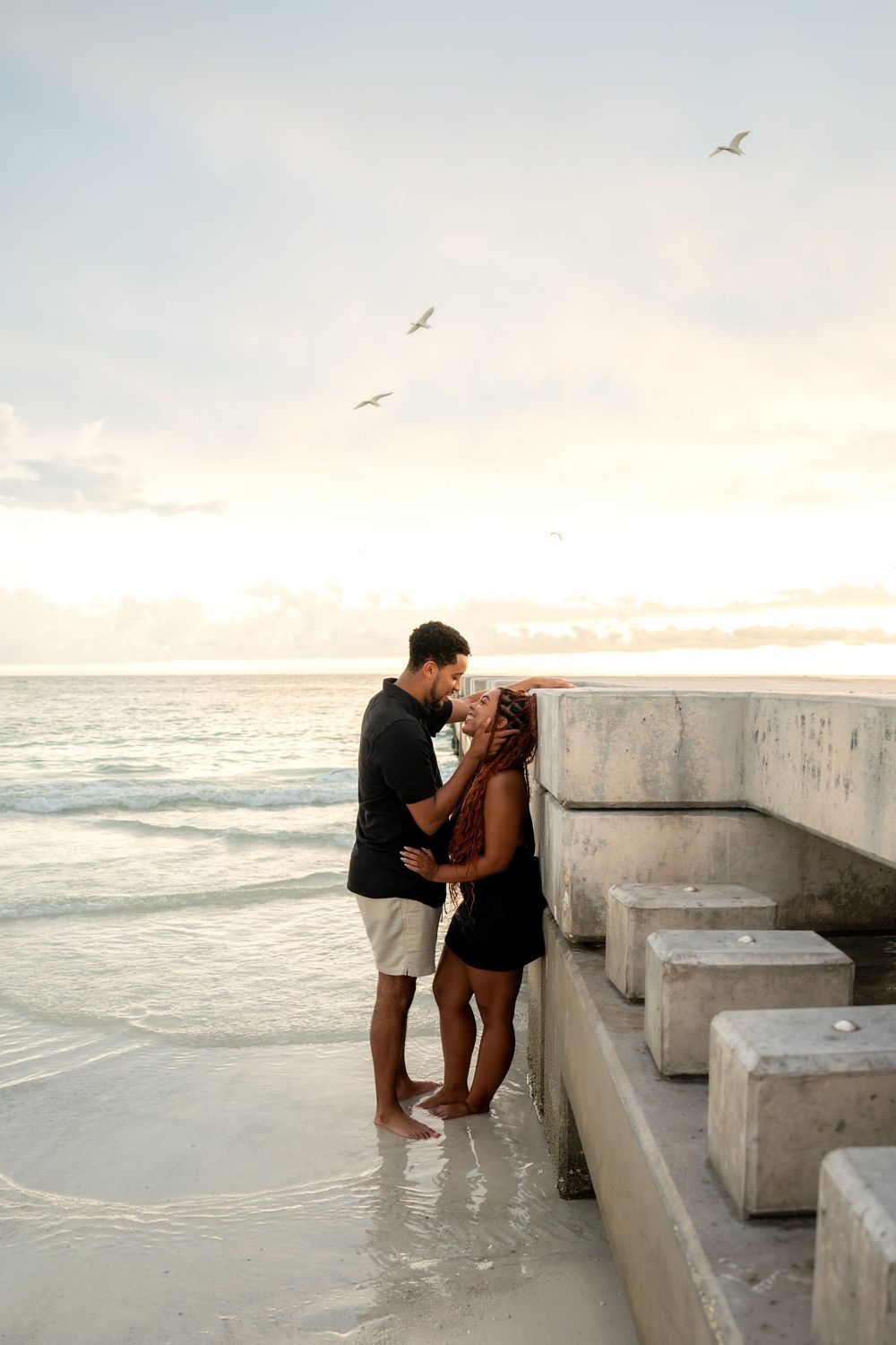 Couple sharing a romantic moment by concrete steps on a beach at sunset with seagulls flying overhead.