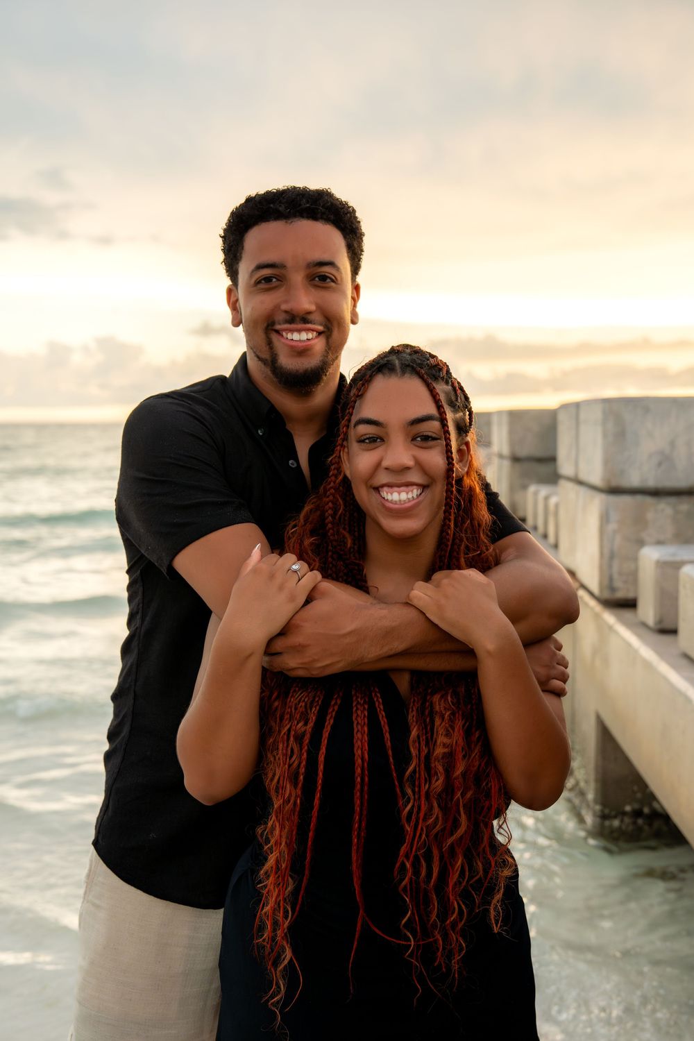 A smiling couple embraces on a seaside pier at sunset, the woman with red braids and the man in a black shirt.