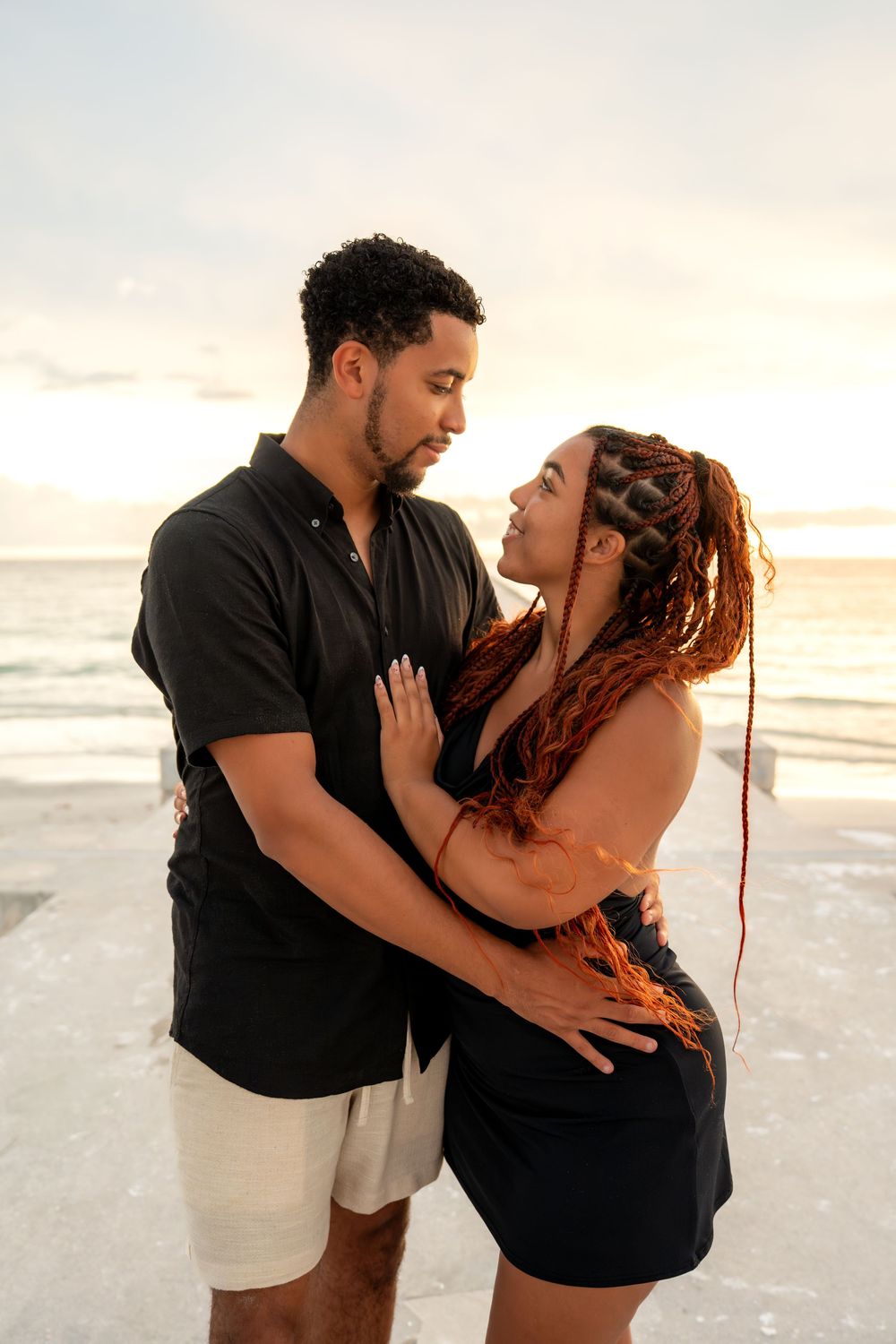 Couple embracing romantically on a beach at sunset, woman with braided hair in black dress, man in black shirt and white shorts.