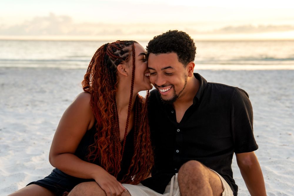 Couple sitting on beach at sunset, woman whispering in mans ear as he smiles joyfully.