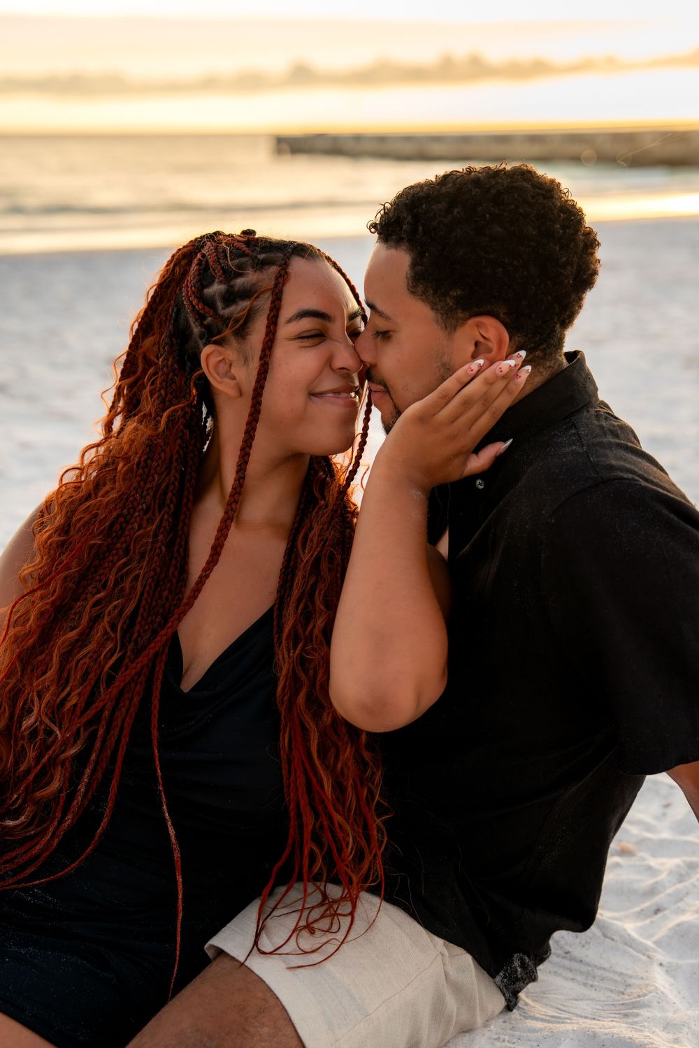 Couple sharing a romantic moment at sunset on the beach, woman with long red braids smiling as man kisses her cheek.