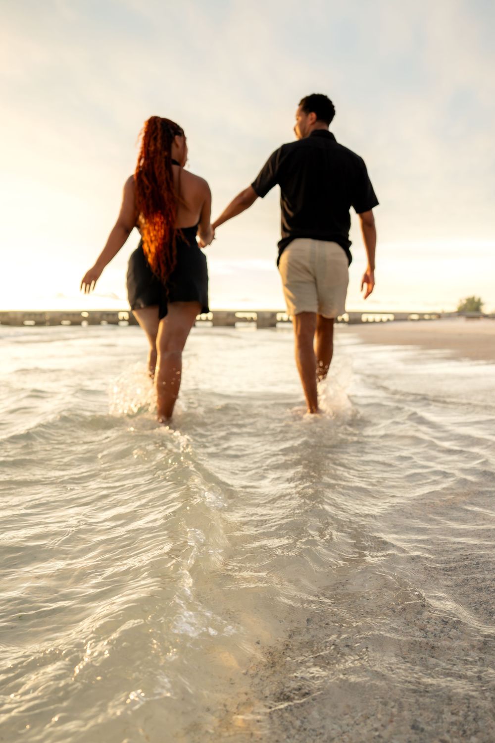 Couple holding hands walking through ocean waves on a sandy beach at sunset.