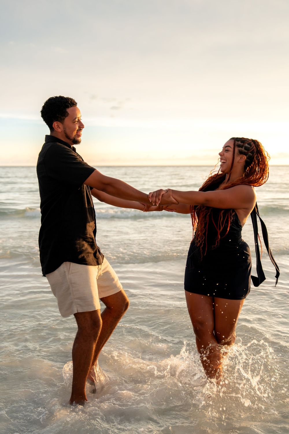Couple holding hands and splashing in ocean waves at sunset, wearing matching black outfits on a tropical beach.