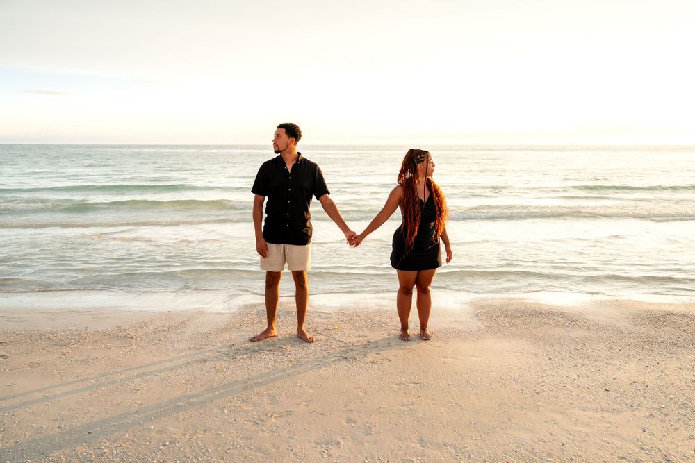 Couple holding hands on a sandy beach at sunset, both dressed in black, with calm ocean waves in the background.