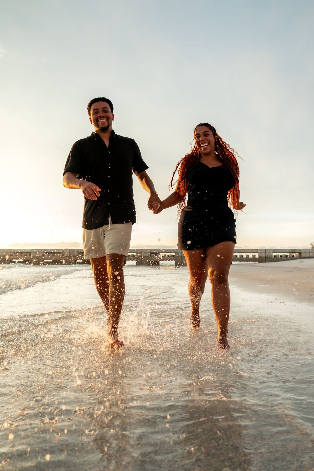 Happy couple running hand-in-hand through shallow ocean waves at sunset, splashing water on a sandy beach.