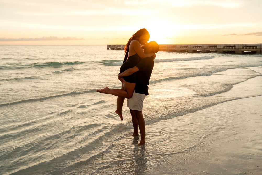 Couple embracing on a sandy beach at golden sunset, waves gently washing ashore near a pier.