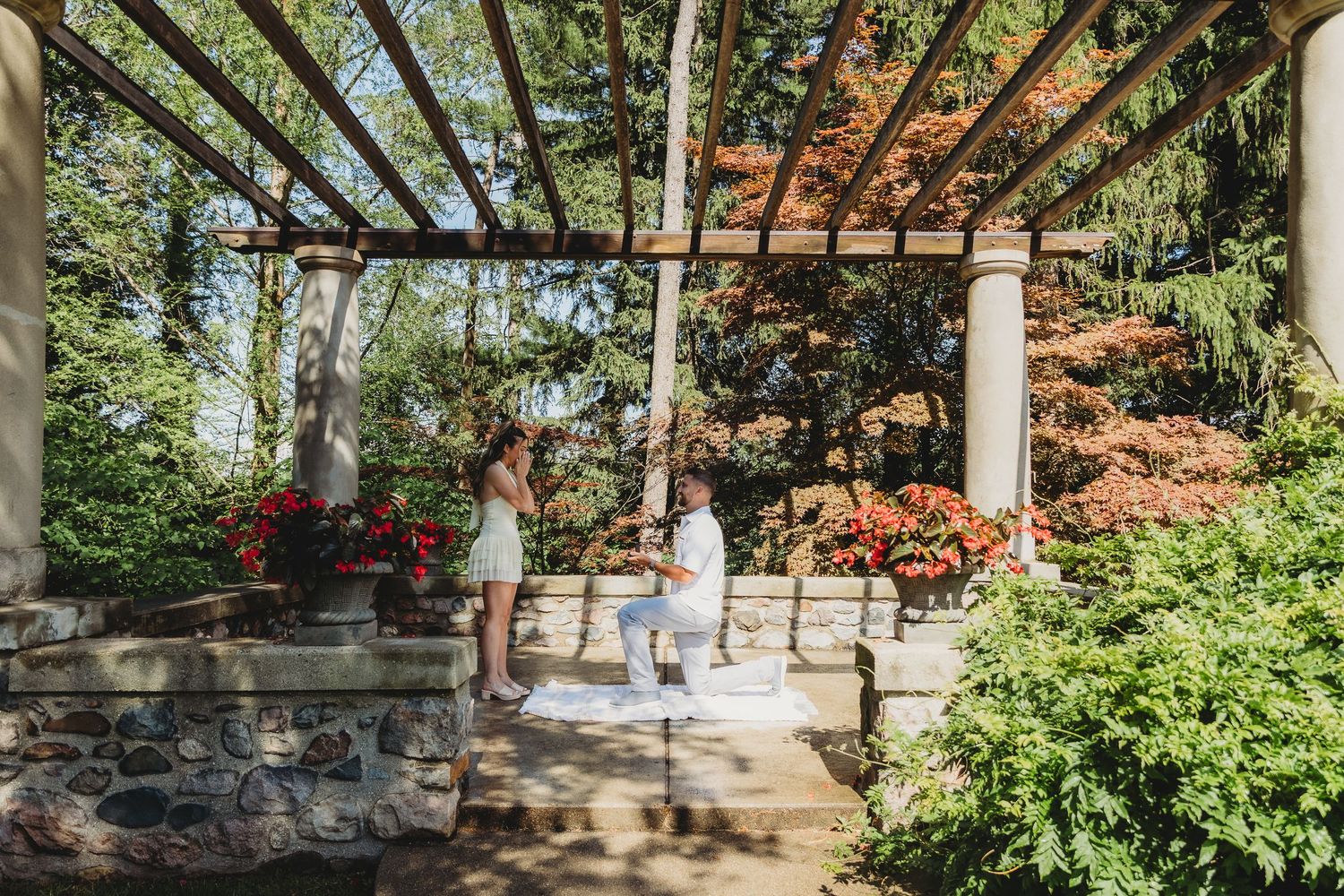 Man proposes on one knee to woman in elegant garden pergola surrounded by colorful flowers and lush greenery.