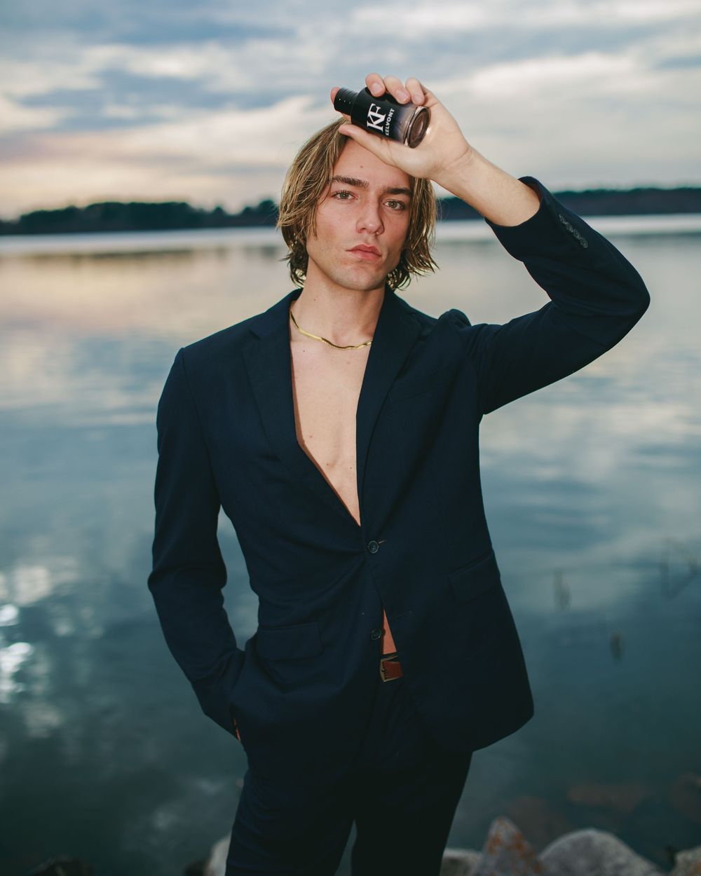 Young man in navy blazer holds fragrance bottle near water at dusk, wearing gold chain necklace.