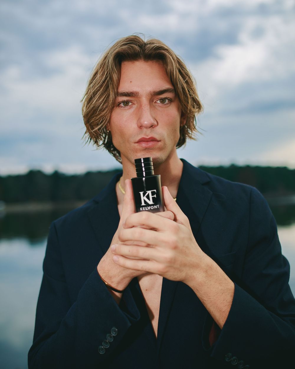 Young man with blonde hair holding KF cologne bottle outdoors near lake wearing navy blazer.