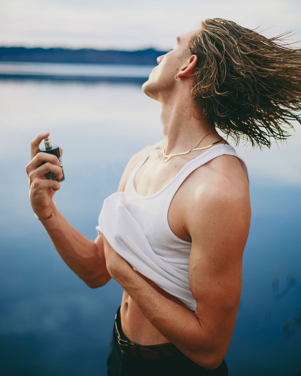 Muscular man in white tank top spraying cologne outdoors by a lake, hair blowing in the wind.
