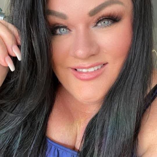 A woman with long black hair and blue eyes smiles in a selfie, wearing a blue top.