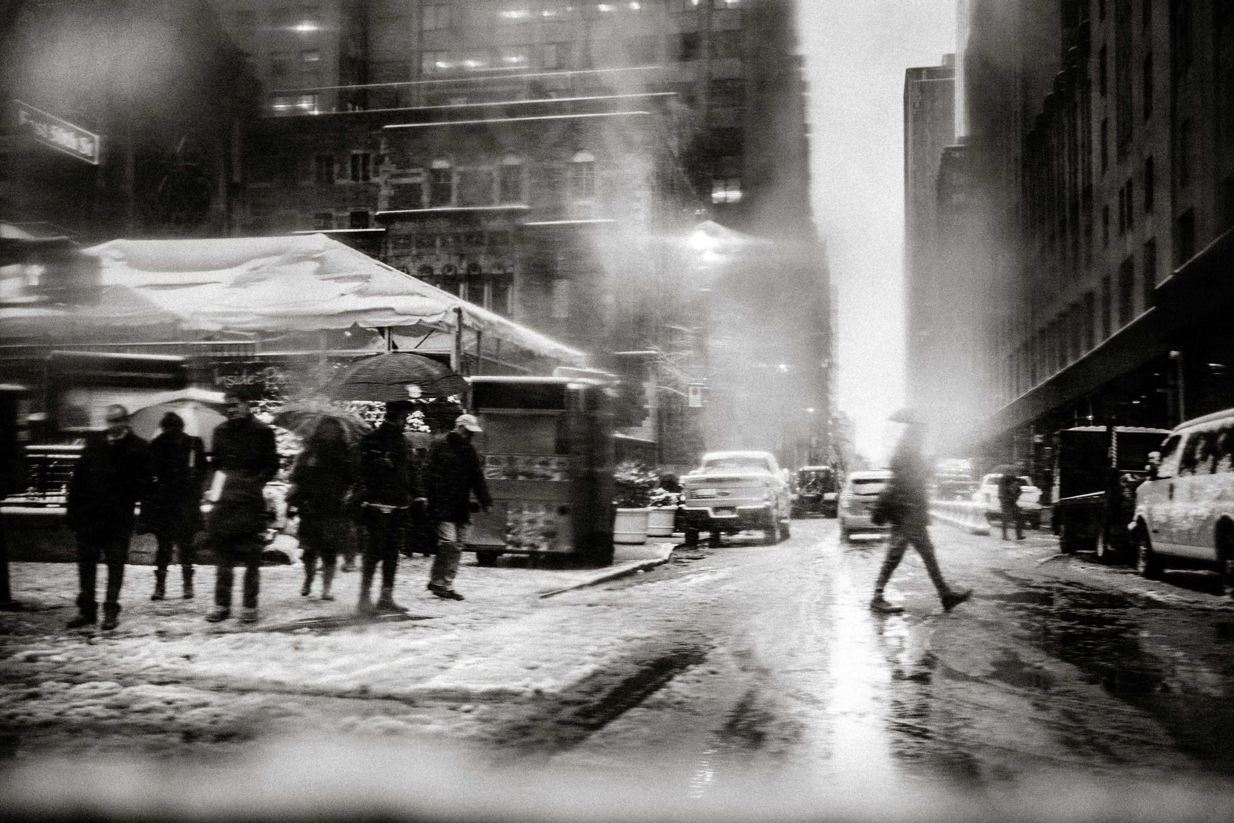 Black and white photo of snowy New York City street with pedestrians and cars amid steam and city lights.