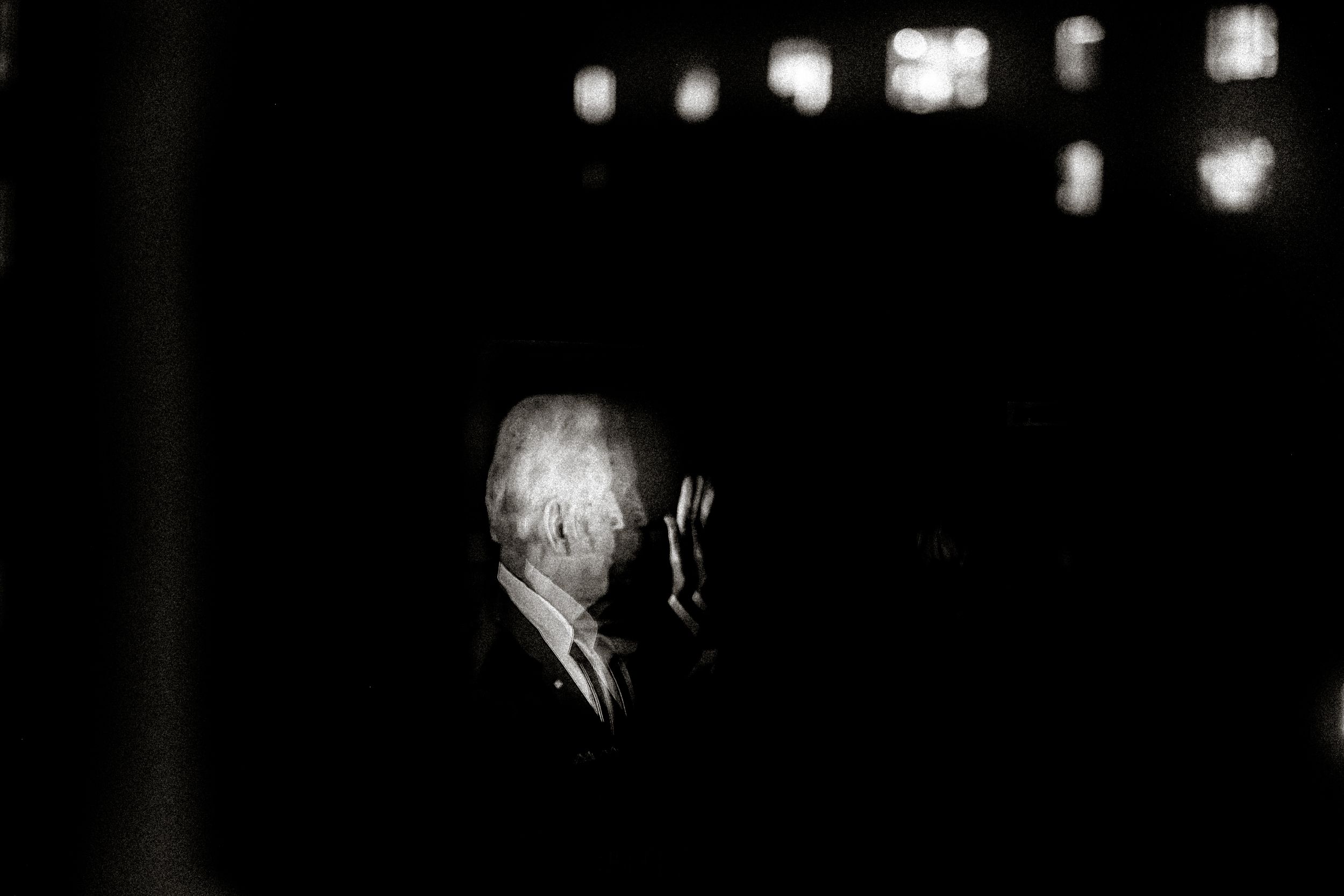 Dramatic black and white portrait of an elderly man in a suit, illuminated against a dark background.