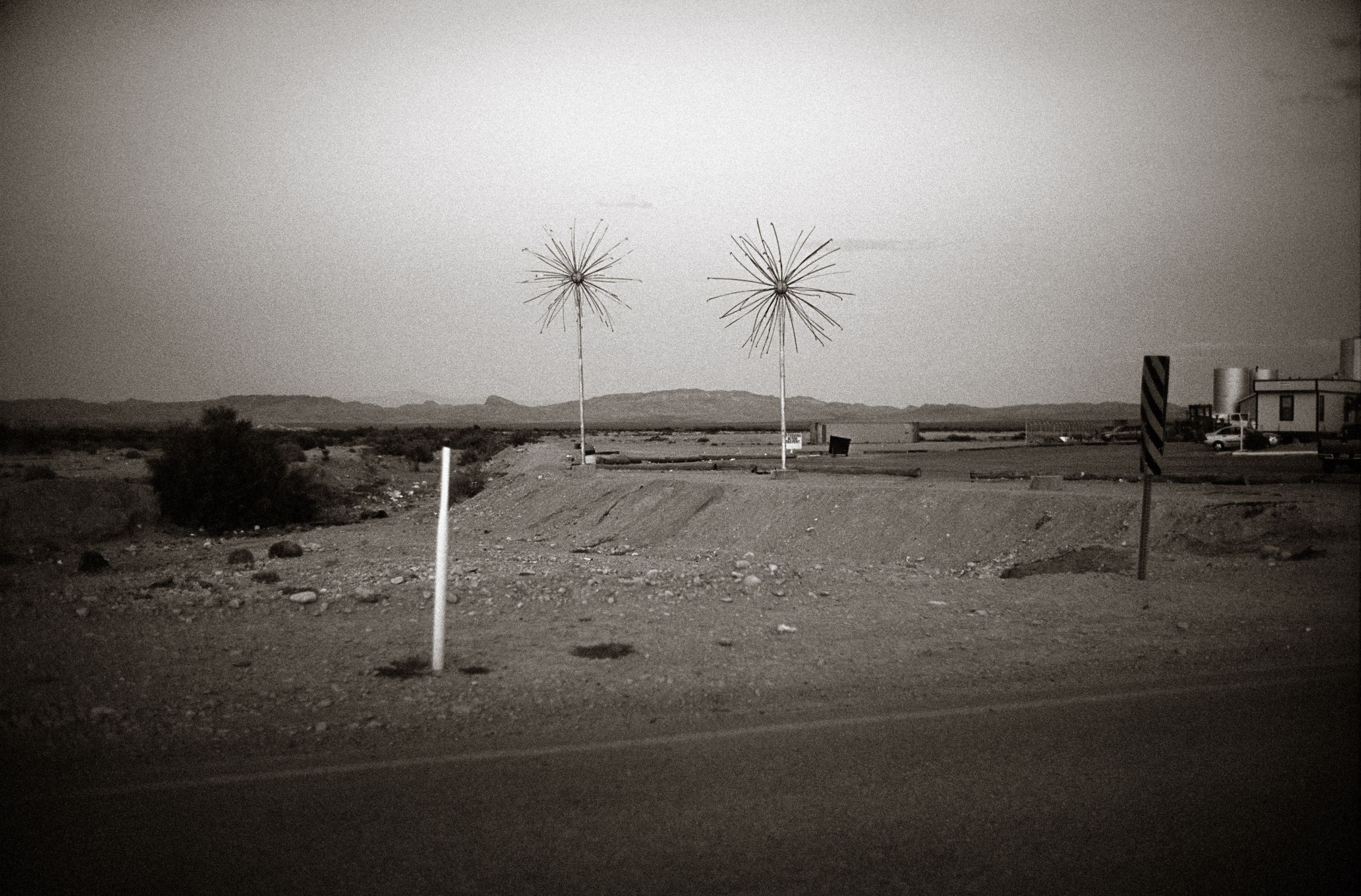 Black and white desert landscape with two starburst sculptures near a roadside building under an overcast sky.