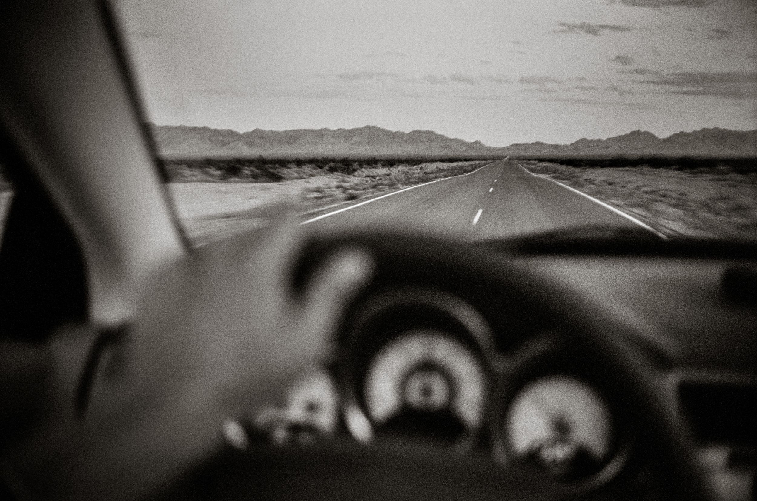 Black and white view from inside a car driving on a long straight desert highway toward distant mountains.