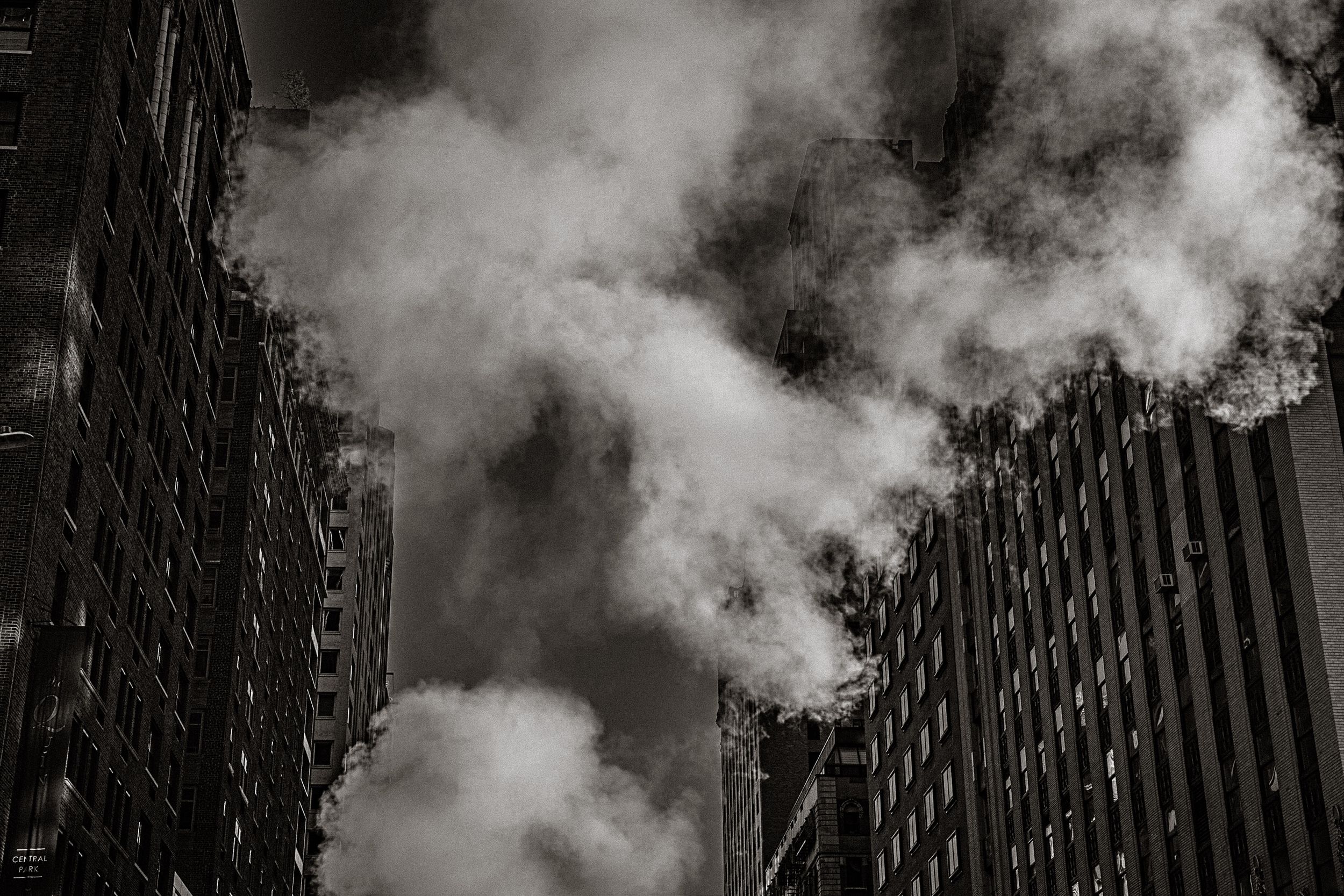 Black and white photo of billowing steam clouds rising between tall city skyscrapers in an urban setting.
