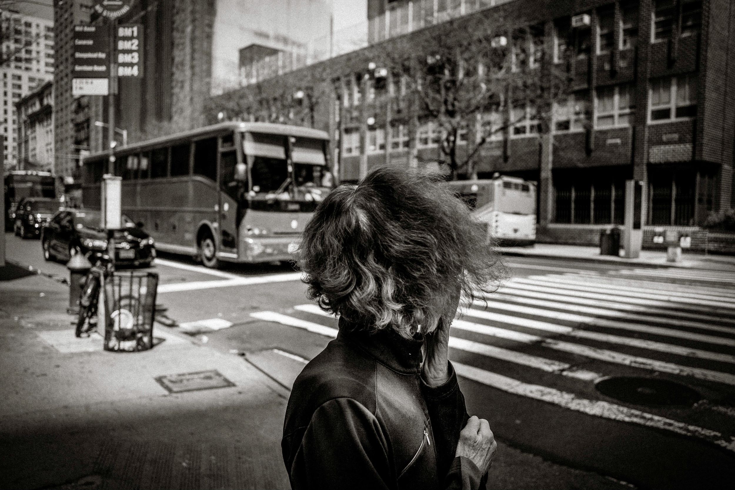 Black and white street photo of a woman with windswept hair on a busy urban intersection with buses and buildings.