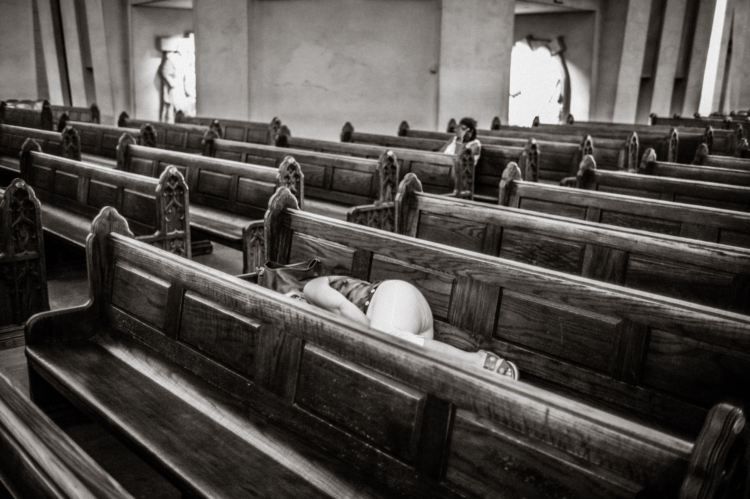 Black and white photo of a bride lying on wooden church pews in a historic chapel interior.