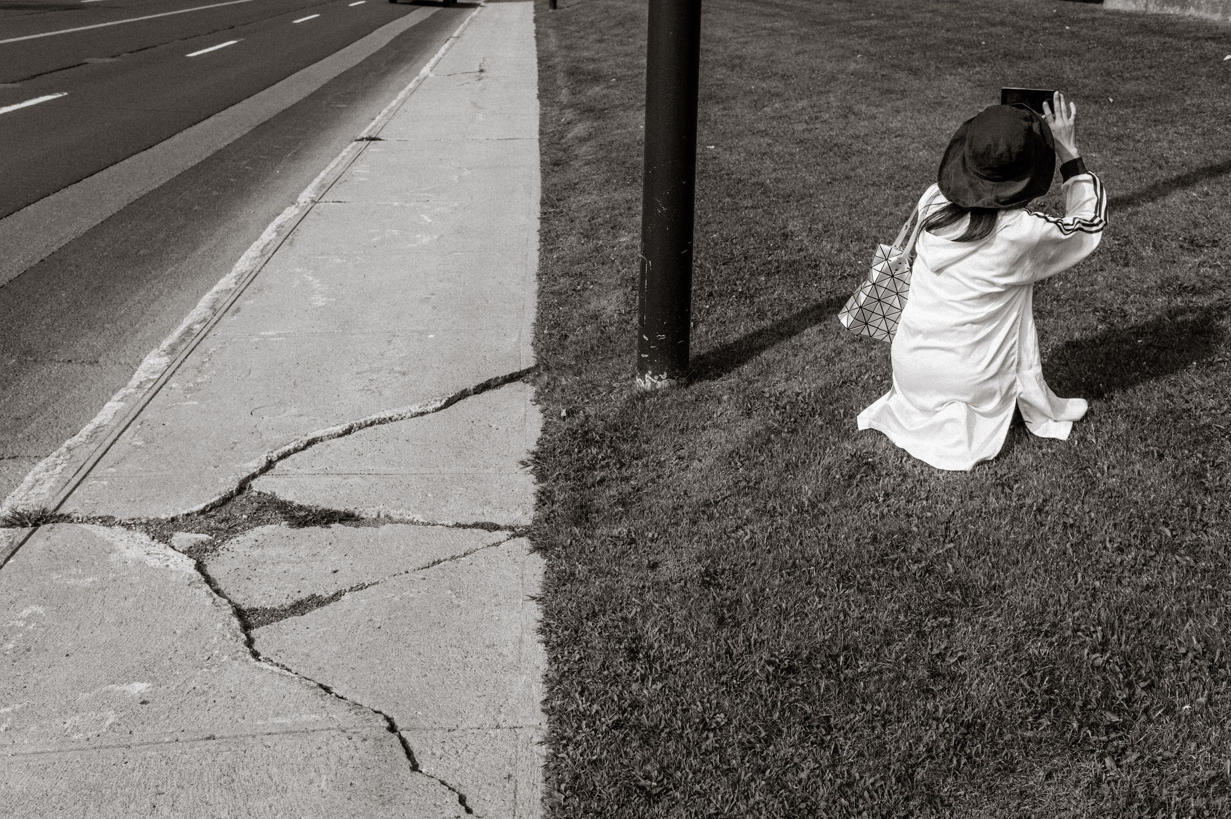 Black and white photo of a child kneeling on grass near a cracked sidewalk and utility pole beside a road.
