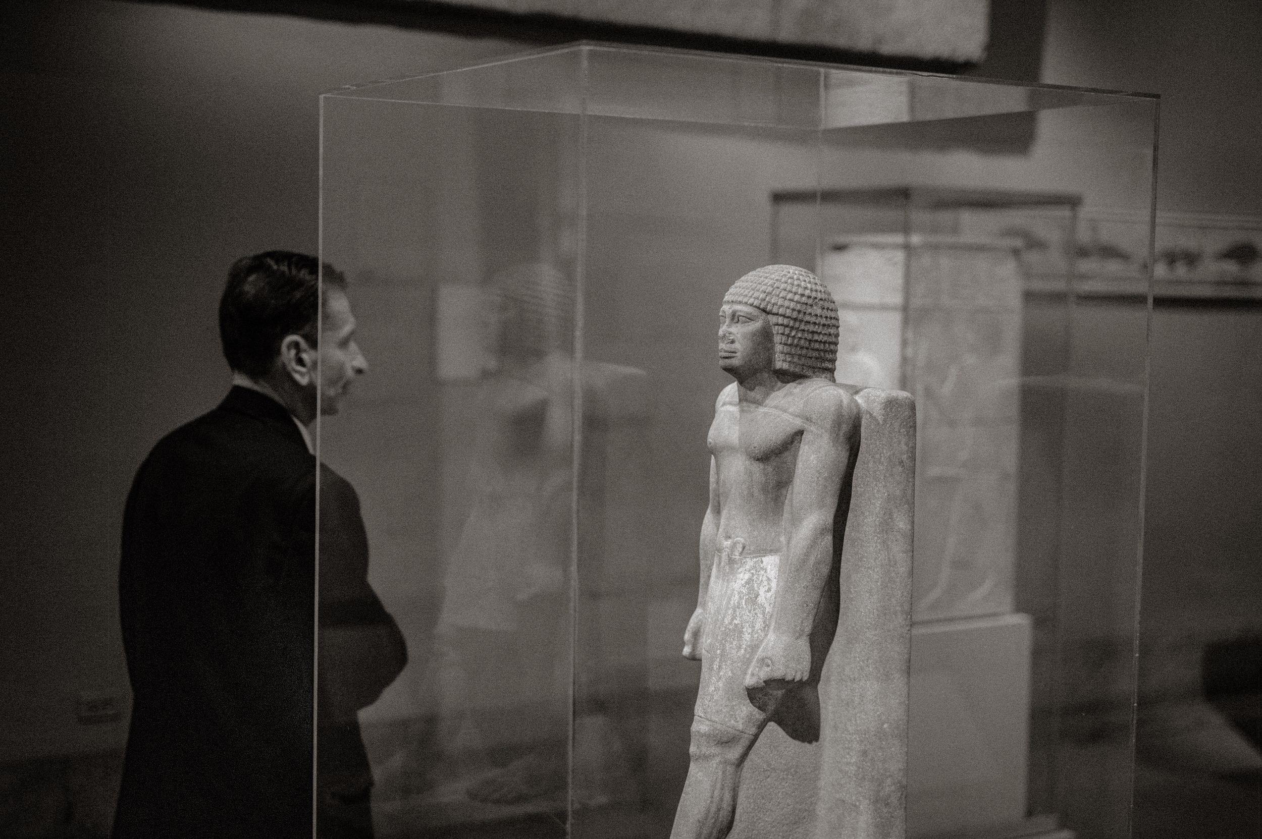 Man in black jacket examining ancient Egyptian statue in glass case at museum, black and white photo.