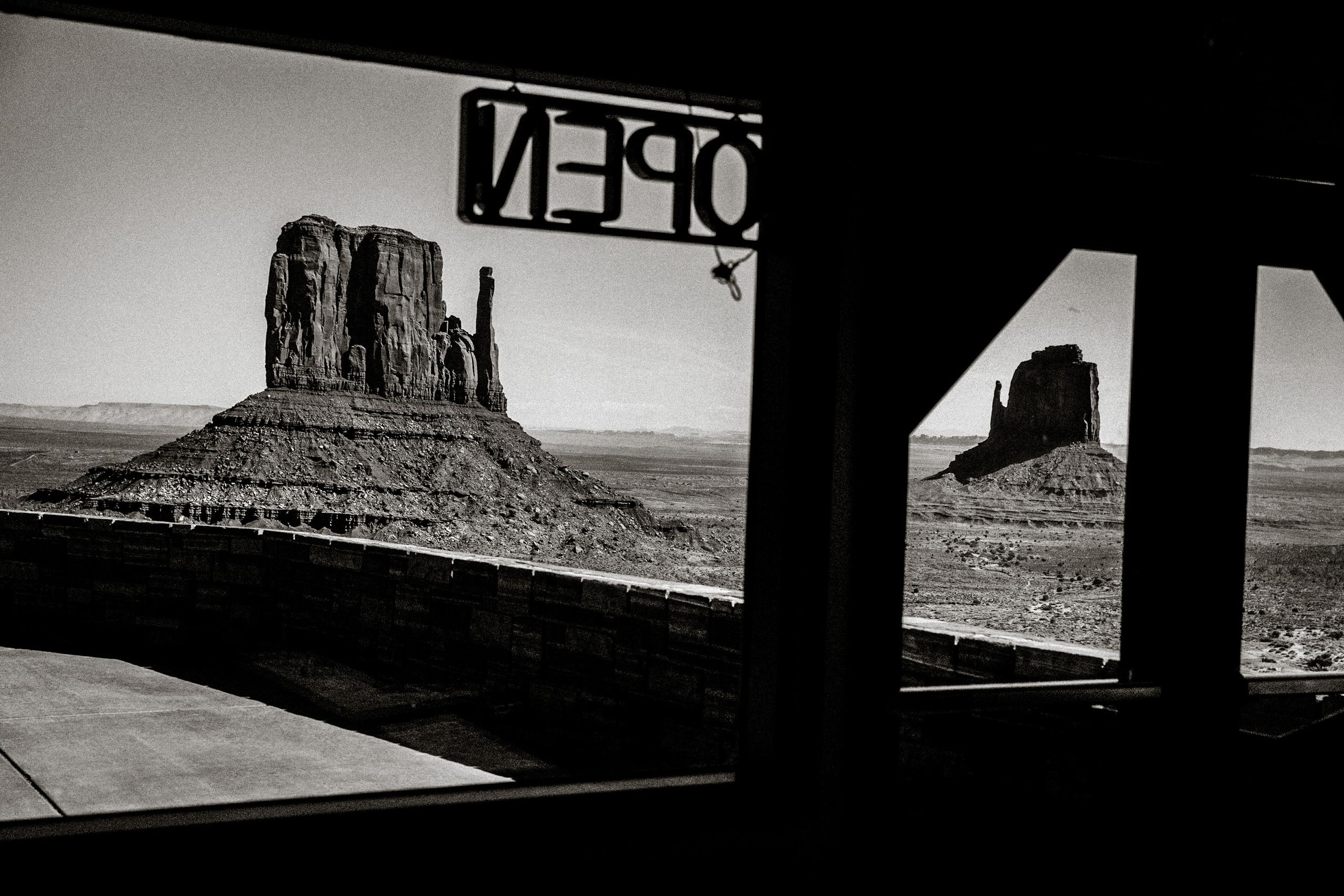 Black and white photo of Monument Valley buttes framed by a building entrance with an OPEN sign in Arizona desert.