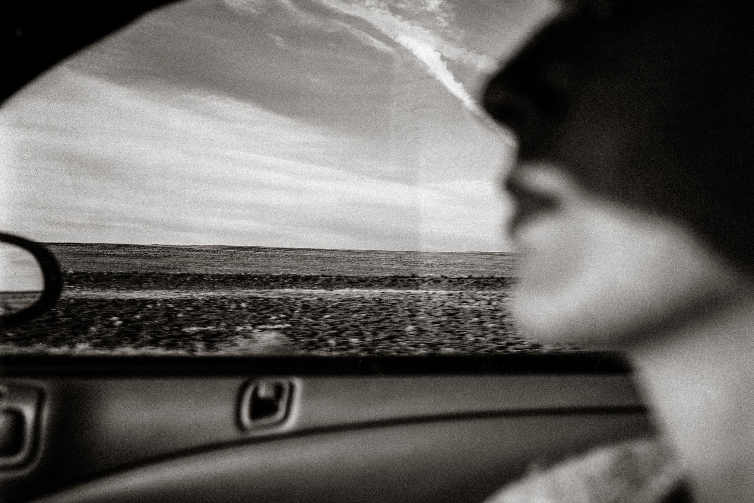 Black and white photo of a person in a car gazing out the window at a vast, flat landscape under a cloudy sky.