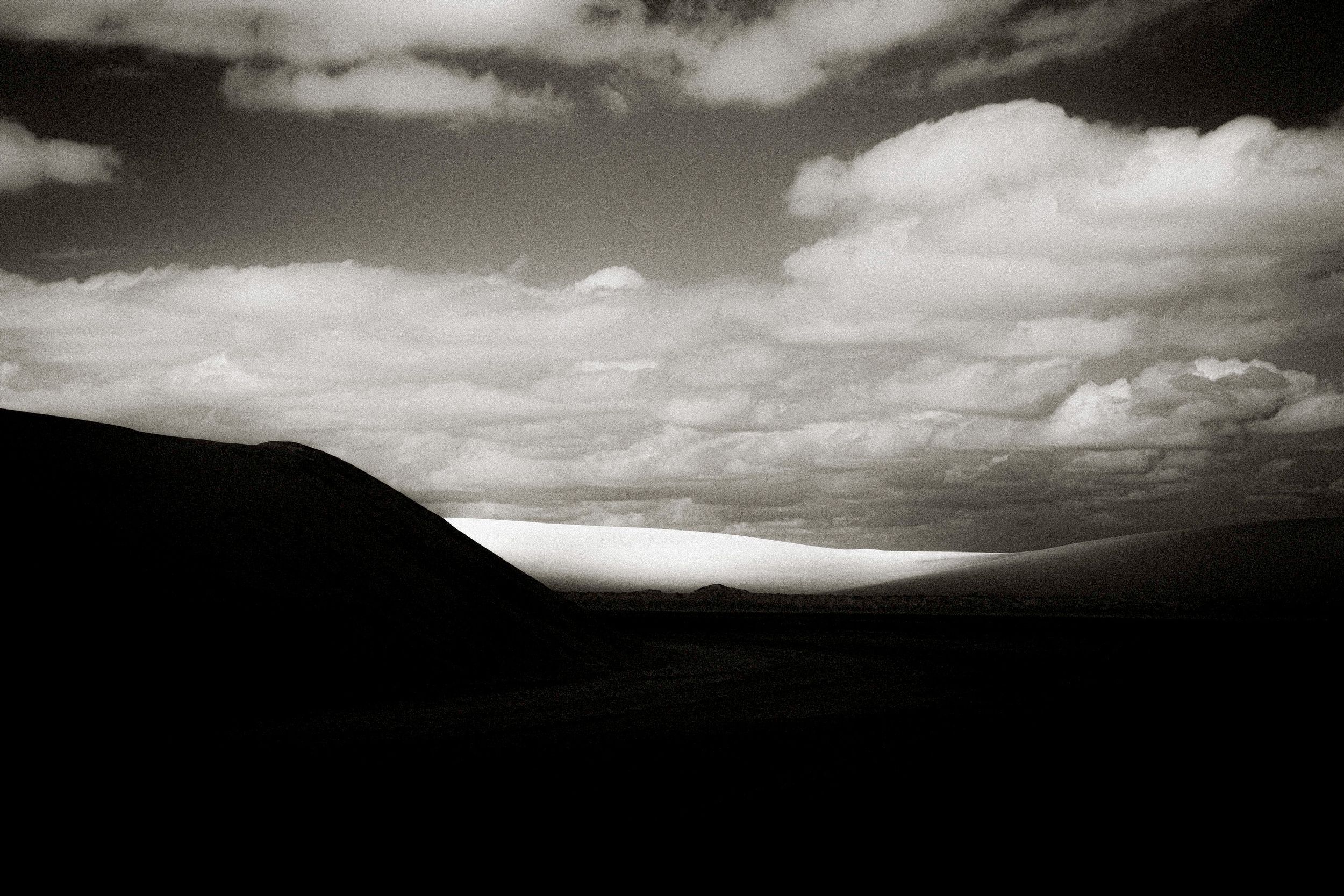 Black and white landscape photo of rolling hills under dramatic cloudy sky with light breaking through on the horizon.