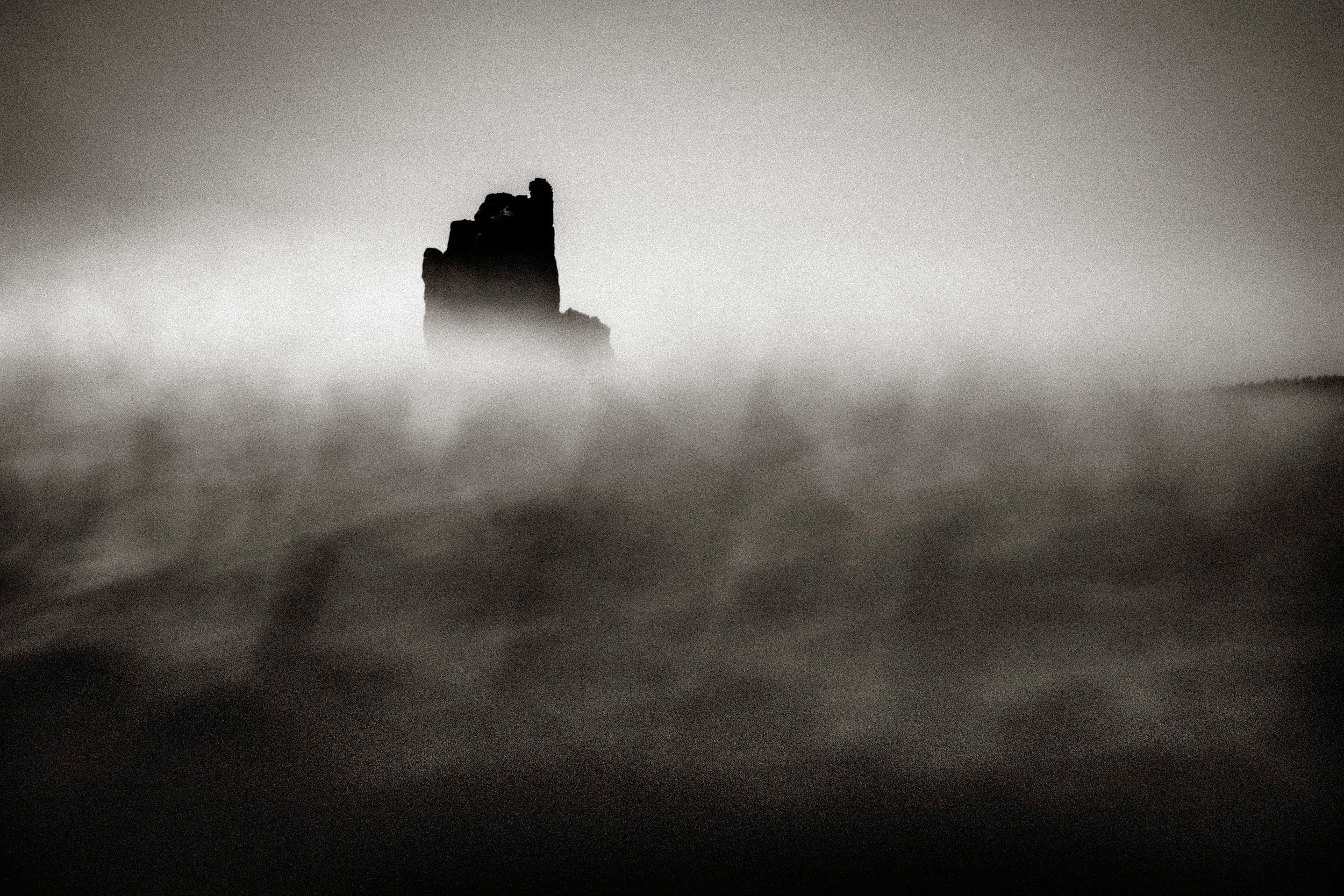 A dark silhouette of a ruined castle emerges through thick swirling fog in a dramatic black and white landscape.