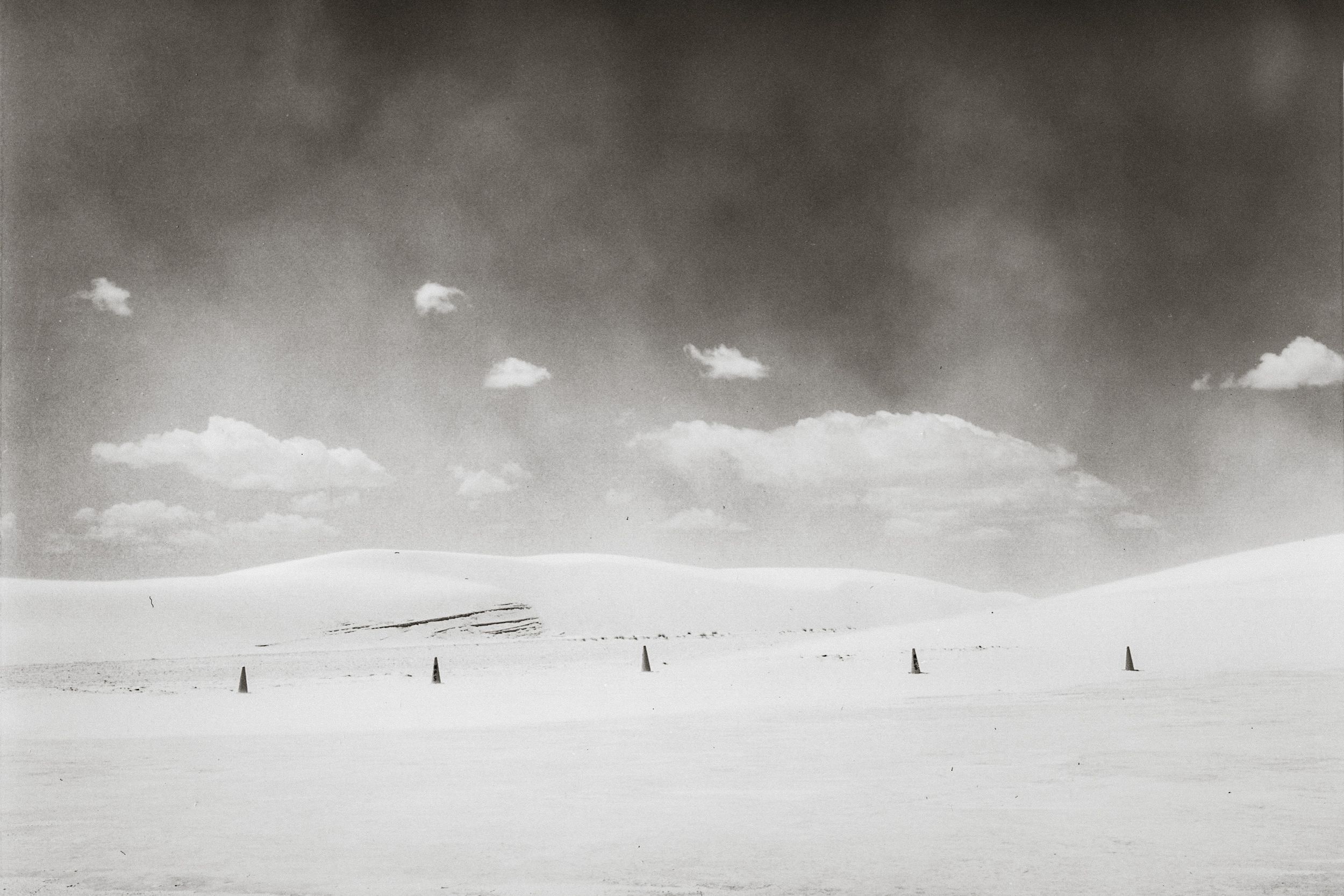 Black and white photo of vast snowy dunes under dramatic cloudy sky with small markers dotting the landscape.