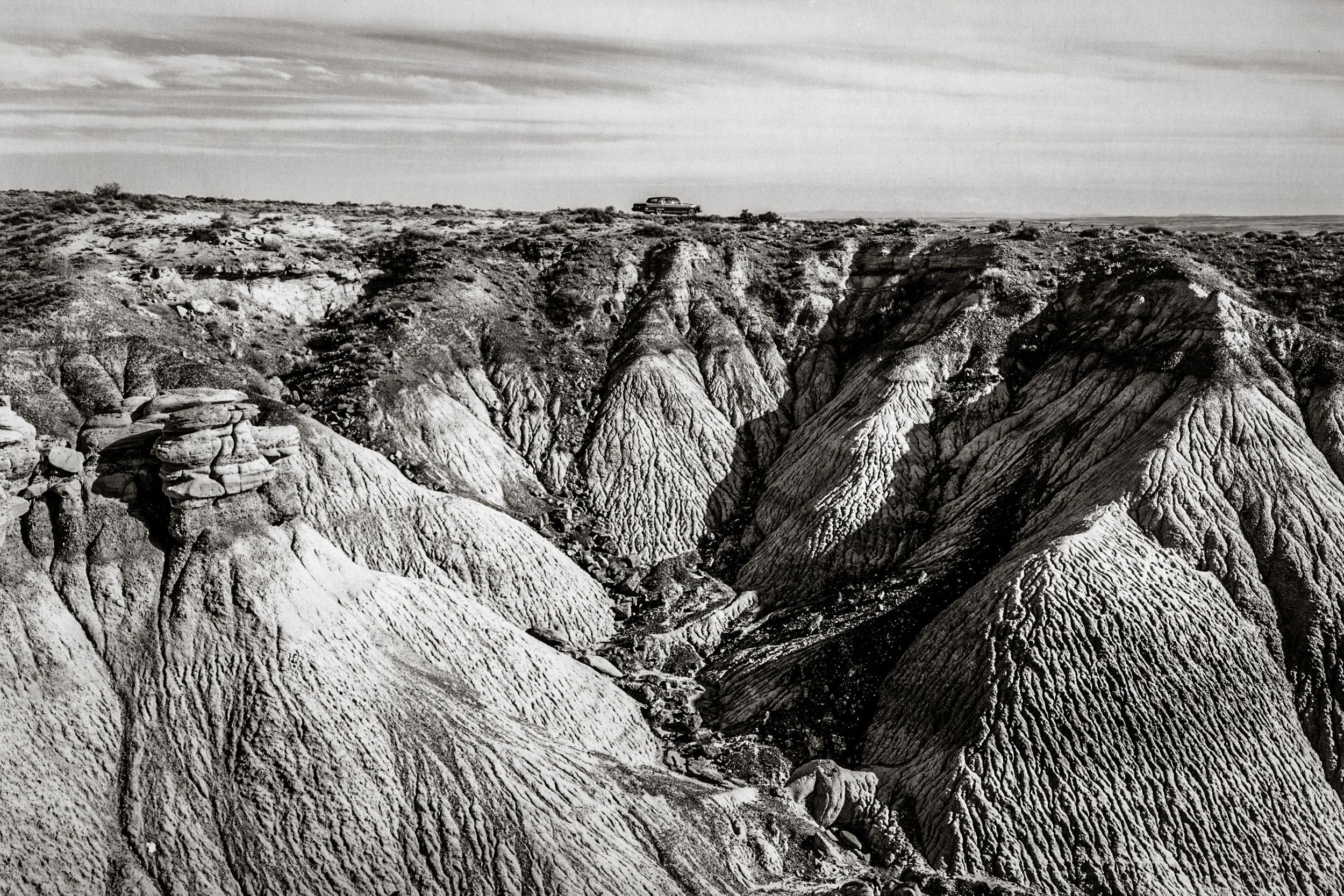 Black and white aerial view of dramatic eroded badlands rock formations with layered ridges under a cloudy sky.