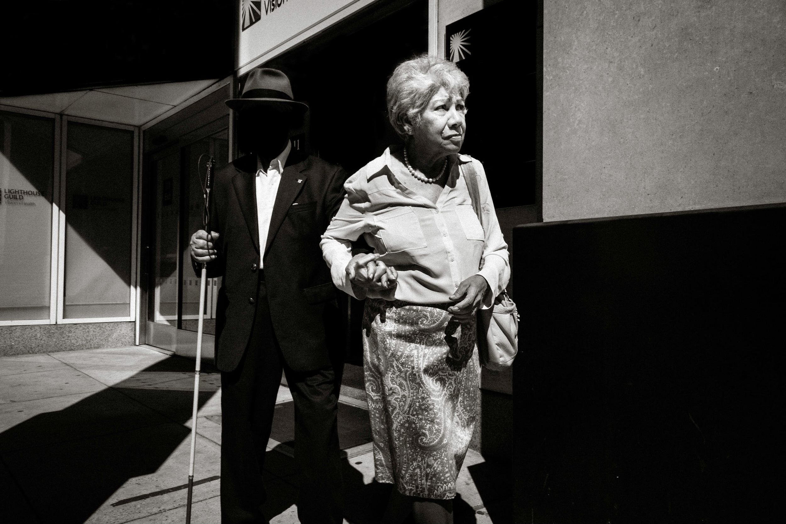 Black and white photo of an elderly couple walking together outside a building, man in hat with cane.