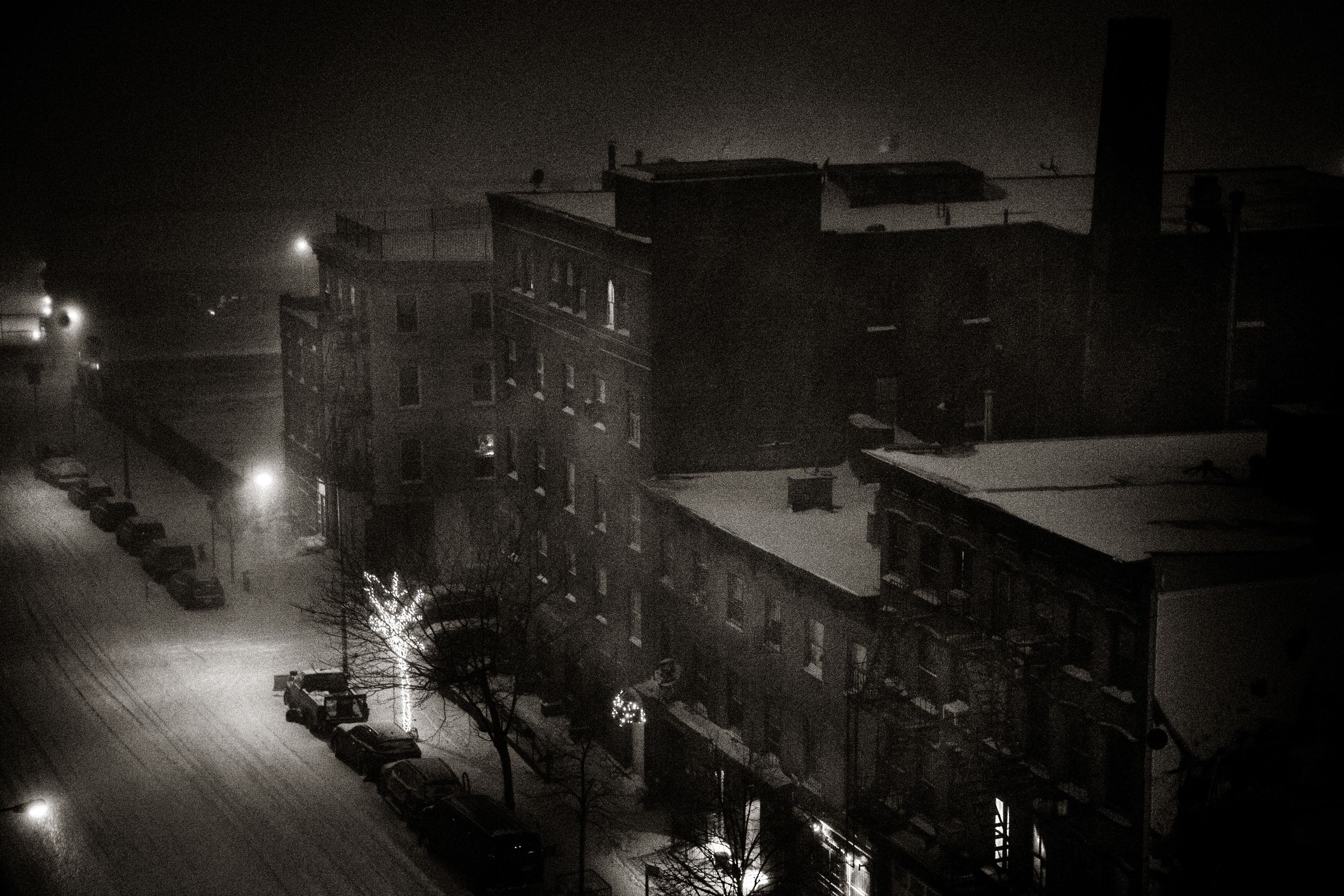 Snowy night cityscape with glowing street lights illuminating snow-covered buildings and a decorated Christmas tree.