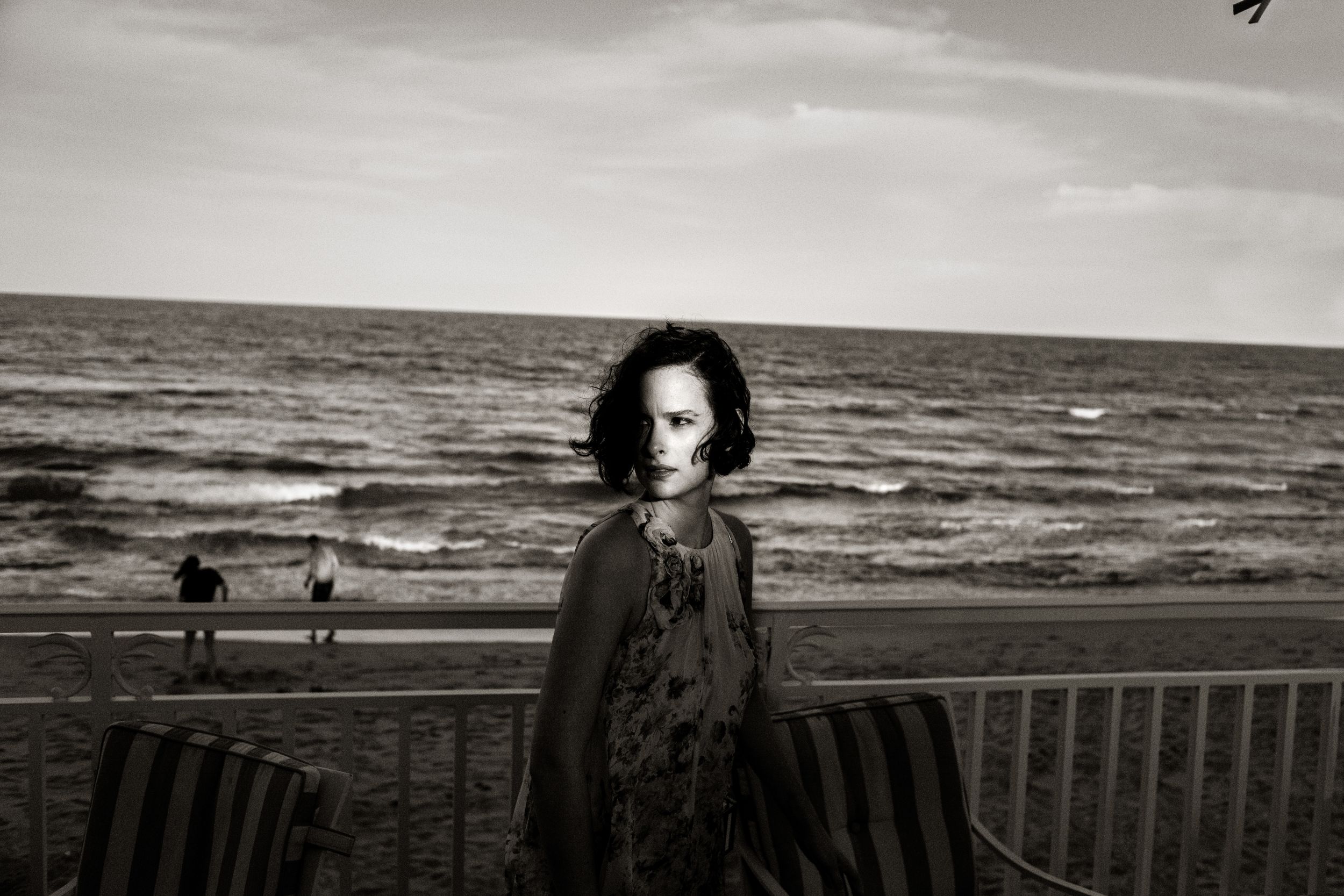 Black and white photo of a woman with dark hair standing on a beach boardwalk with ocean waves in the background.