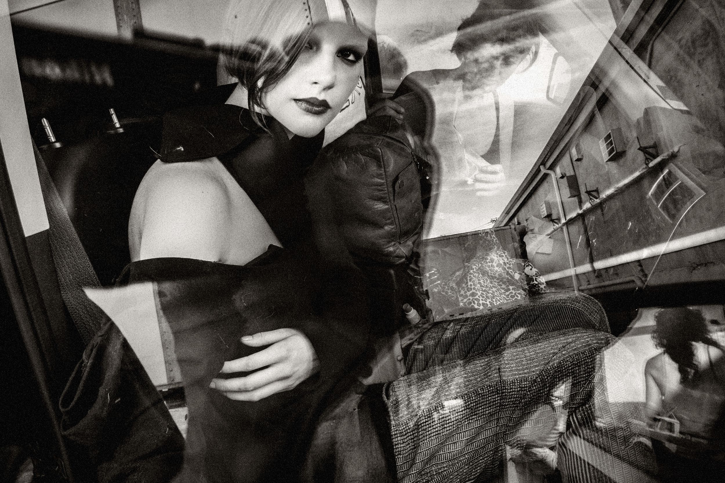 Black and white photo of a glamorous blonde woman in dark clothing, reflected in glass with urban surroundings.