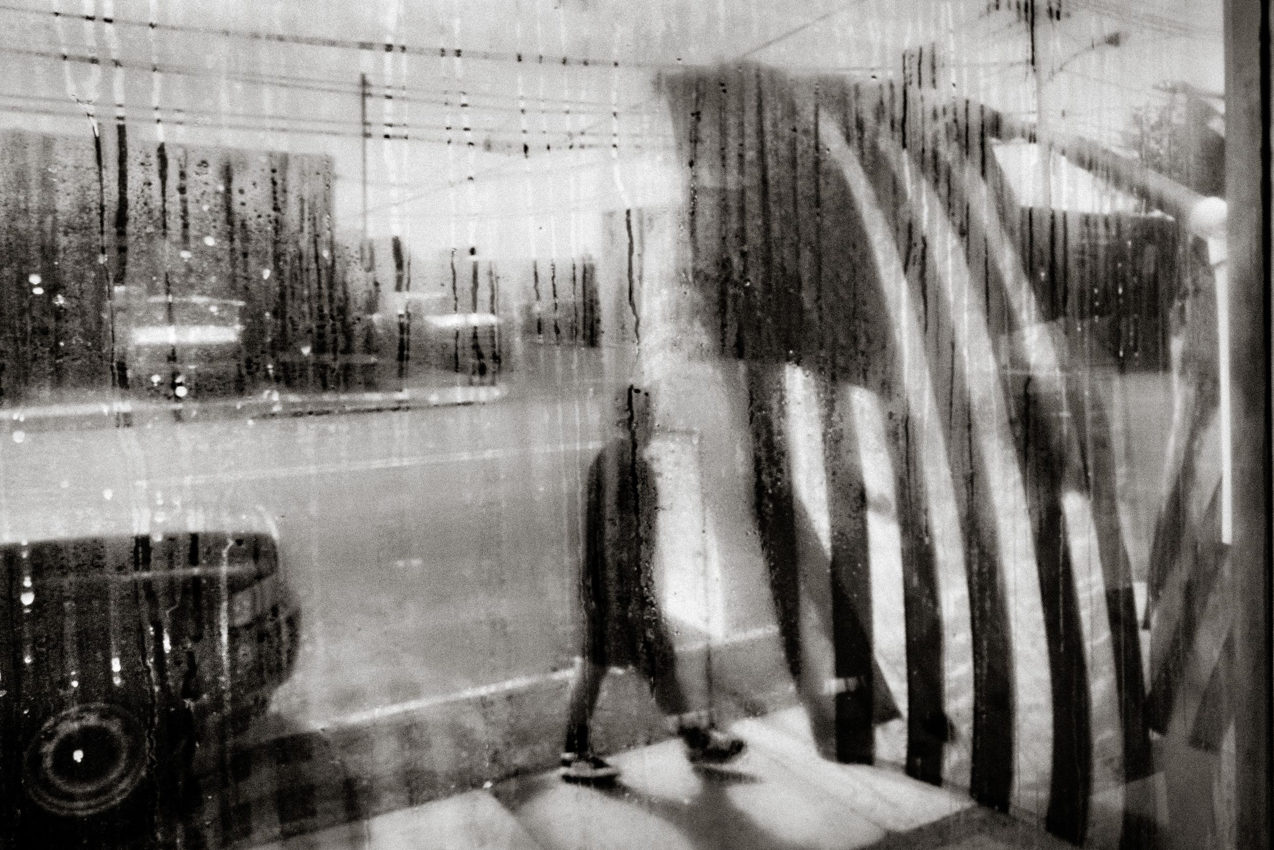 Black and white photo of a rainy day seen through a wet glass window with blurred reflections of a person and flag.