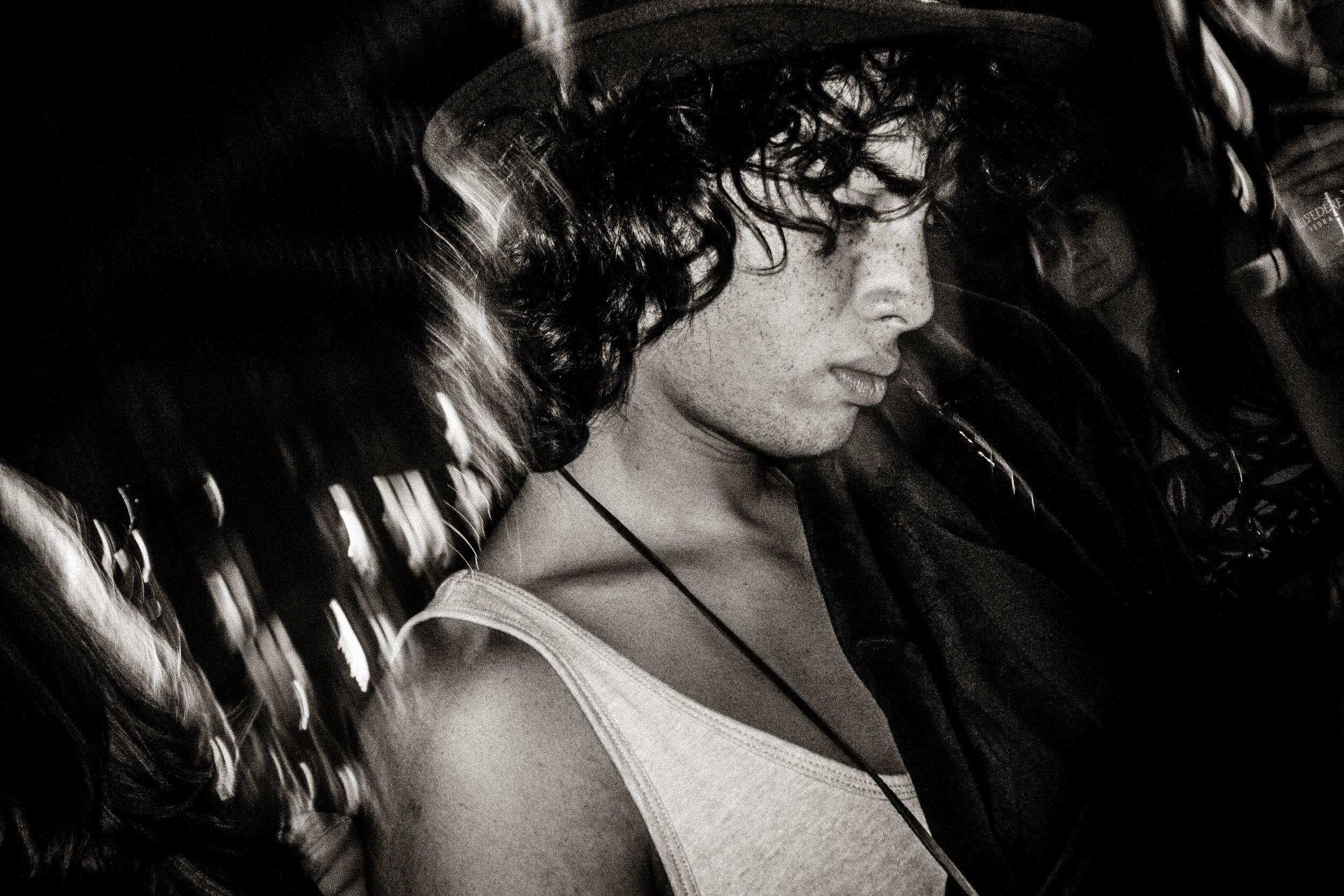 Black and white photo of a young man with curly hair wearing a tank top and necklace in a crowded setting.