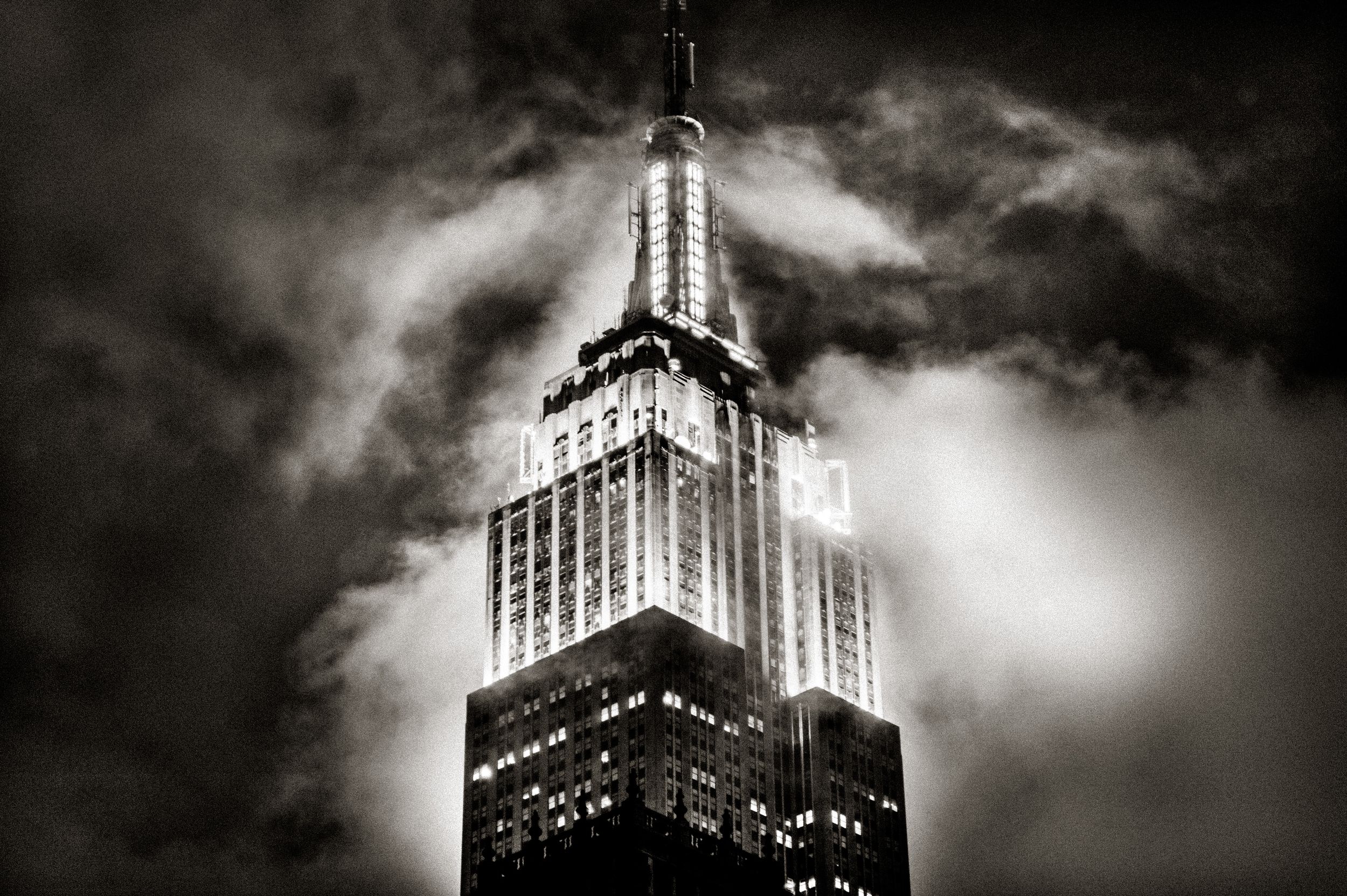 Black and white photo of Empire State Building top shrouded in dramatic storm clouds at night with glowing windows.