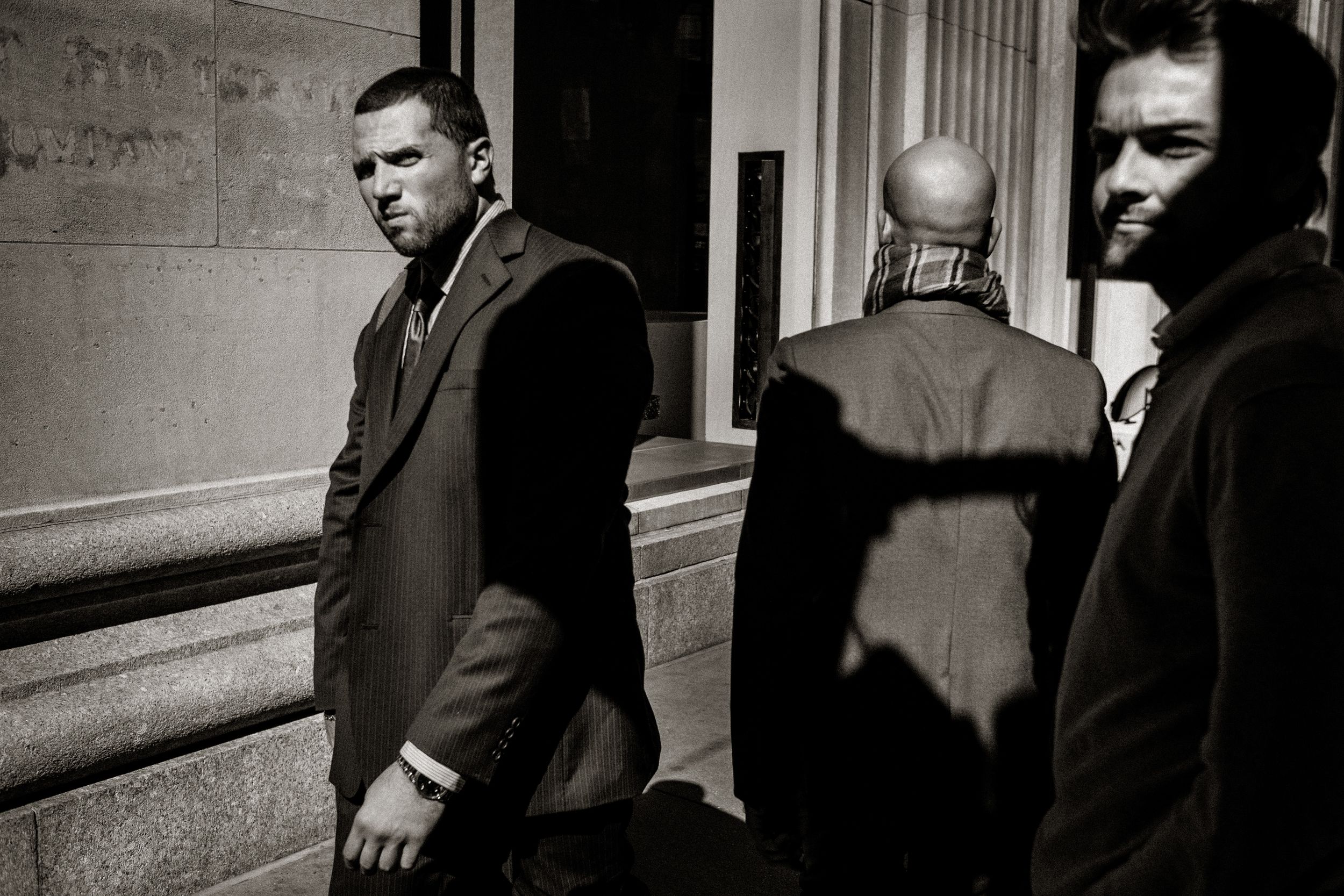 Black and white street photo of three men in suits near a building entrance, captured in dramatic urban light.