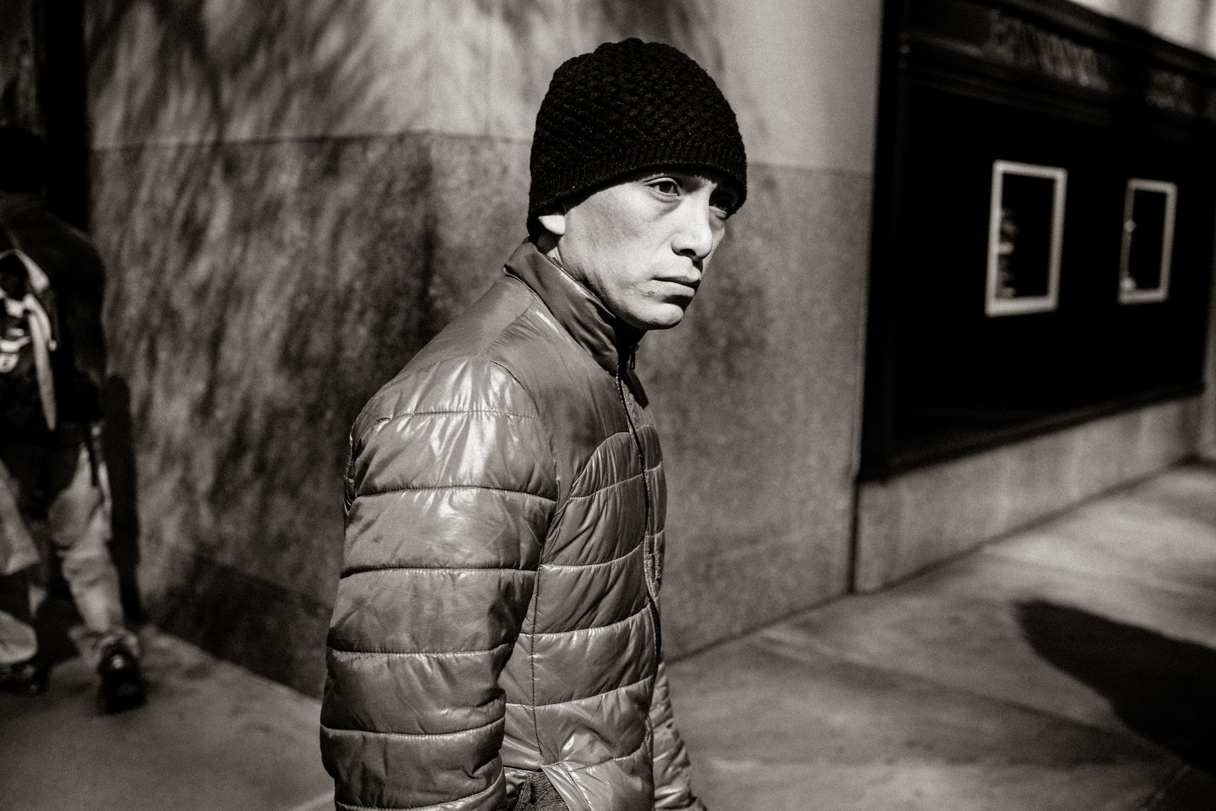 Black and white street photo of a young man in a puffer jacket and beanie walking along an urban sidewalk.