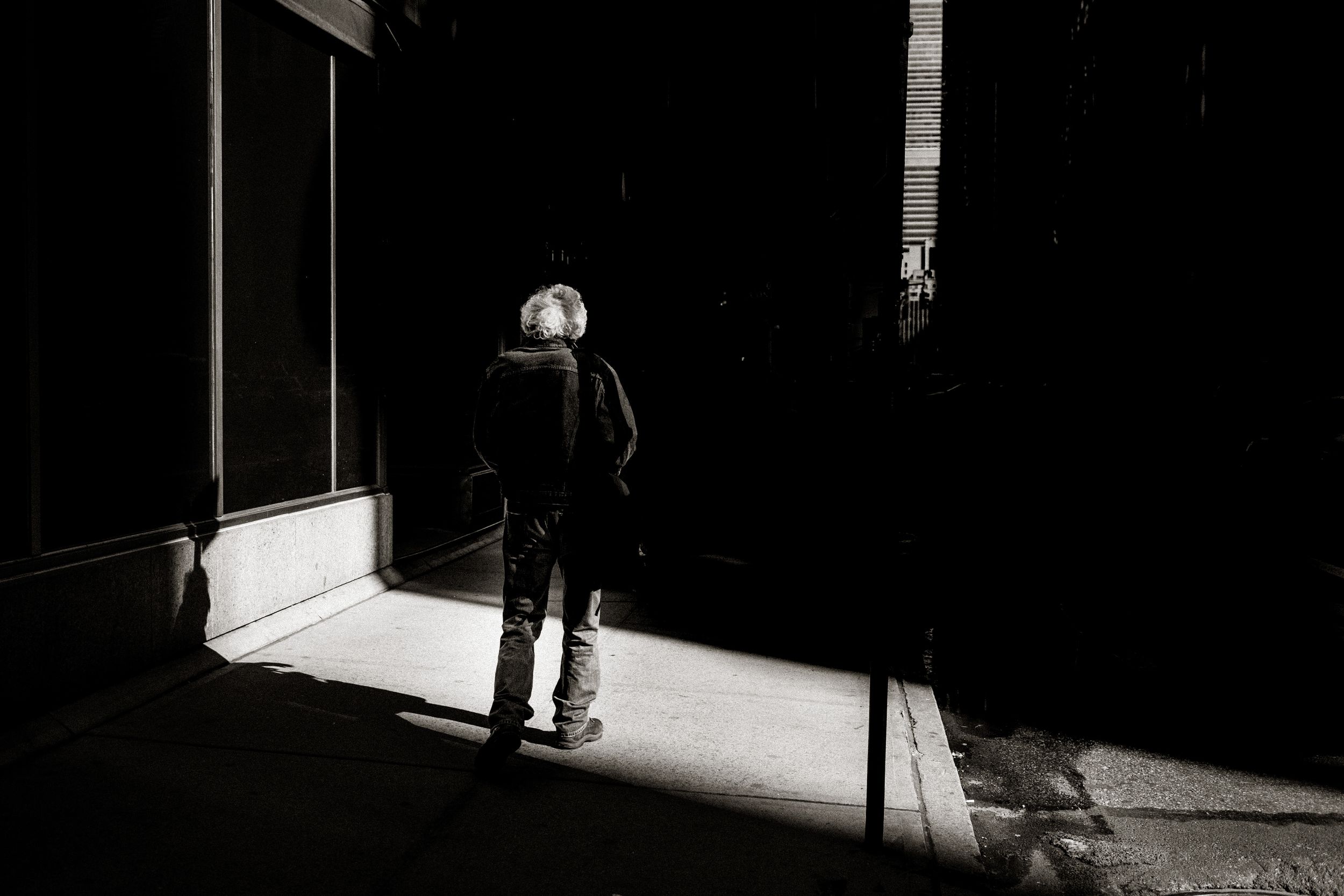 A lone figure stands in a dramatic black and white urban alley, illuminated by a striking shaft of natural light.