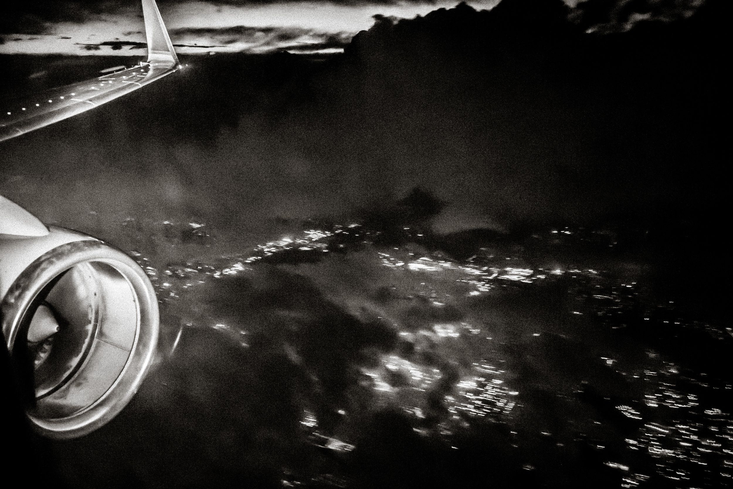 Black and white aerial view from airplane window showing engine and glittering city lights through dramatic clouds at night.